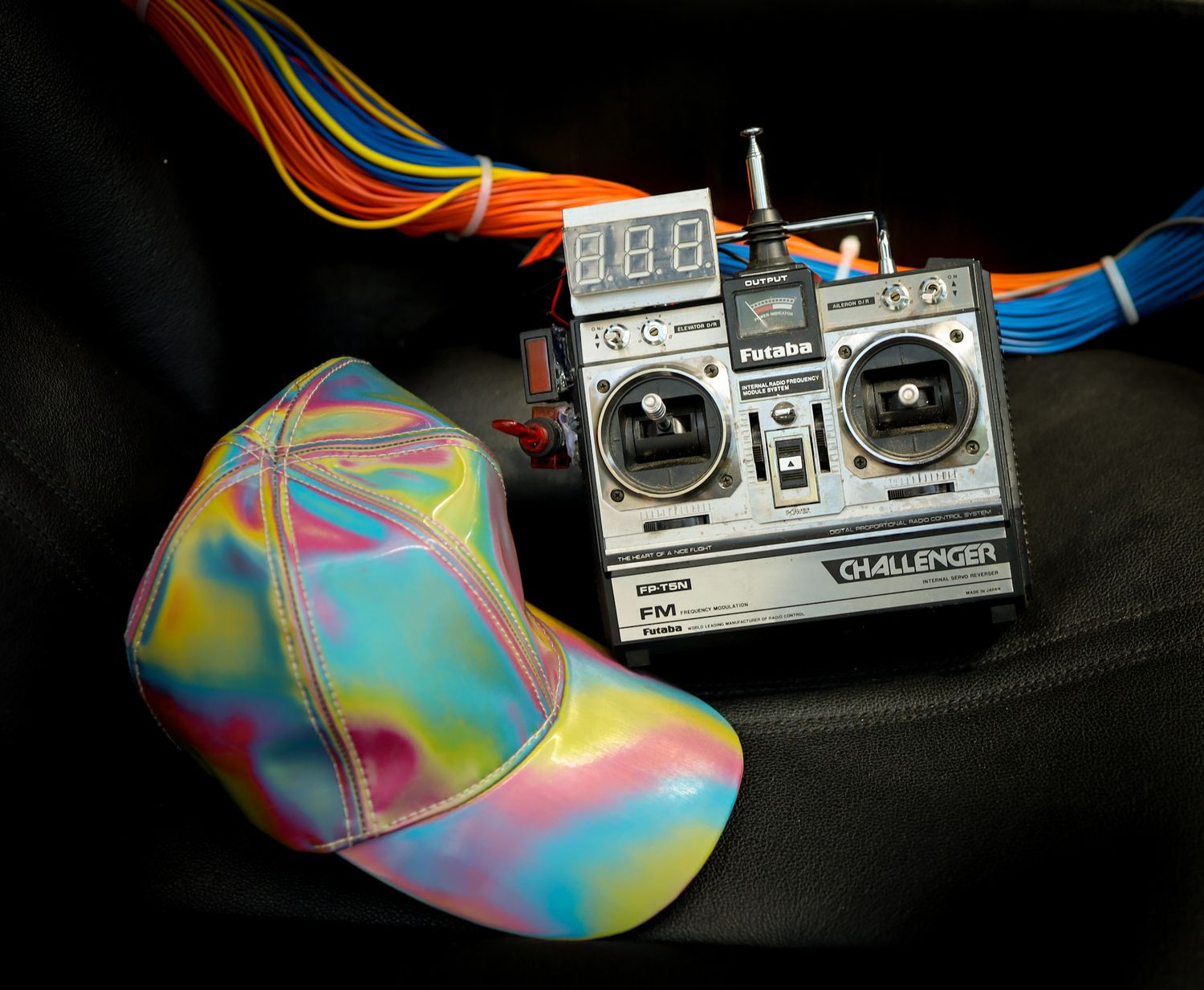 Holographic iridescent baseball cap beside a Futaba Challenger FM RC radio controller on dark background.