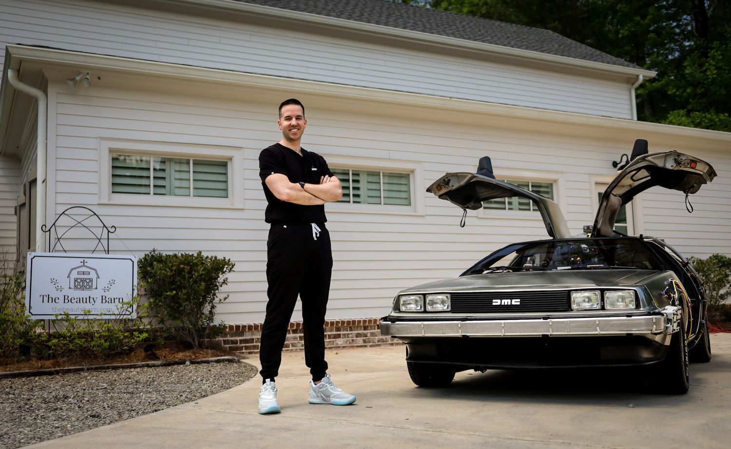 Man in black outfit stands beside a DeLorean DMC-12 with gull-wing doors open in front of a white house.