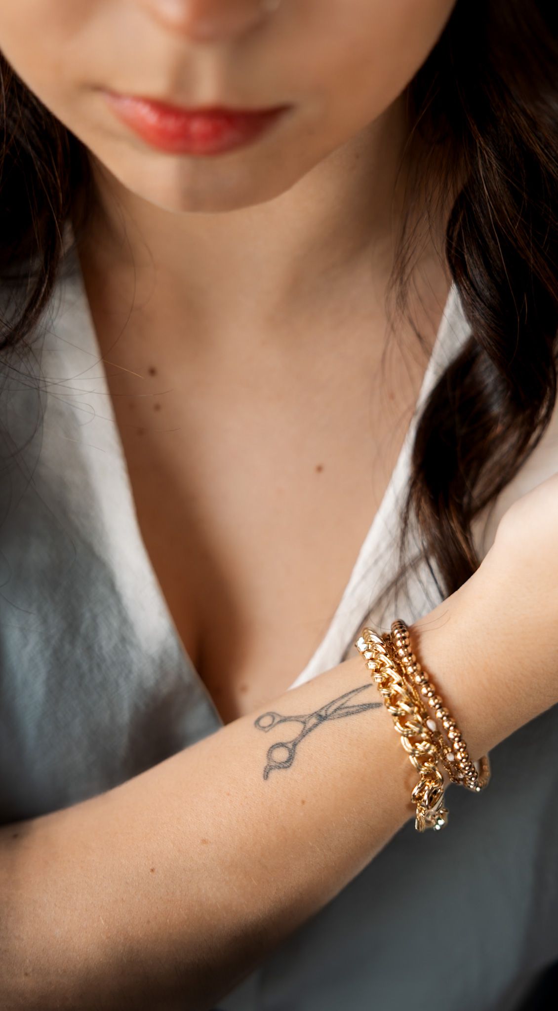 Woman with scissors tattoo on forearm wearing stacked gold chain bracelets, wearing white top with red lipstick.