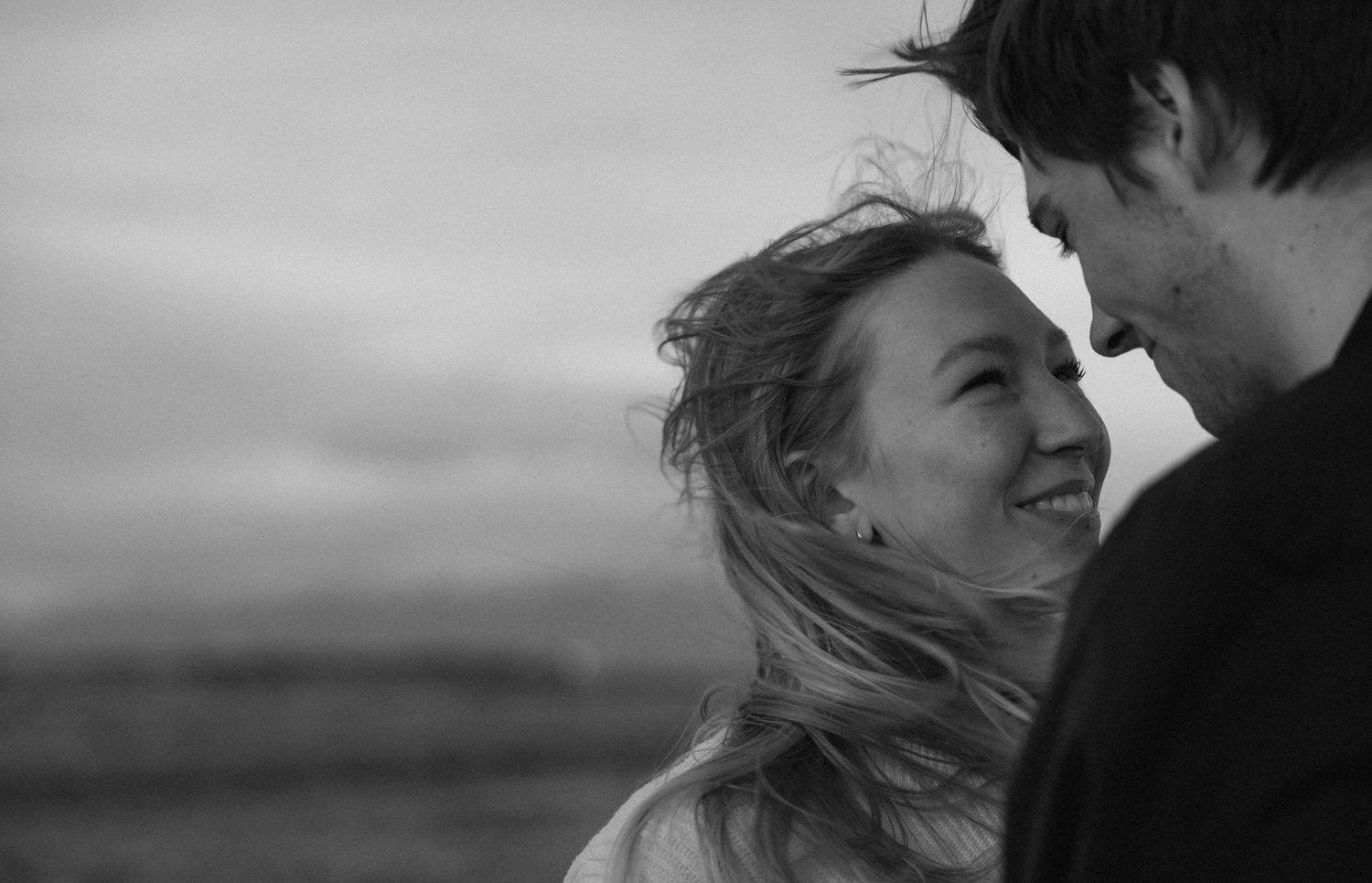a cinematic black and white photo of a couple looking at each other as the wind blows their hair in their faces with the sun set