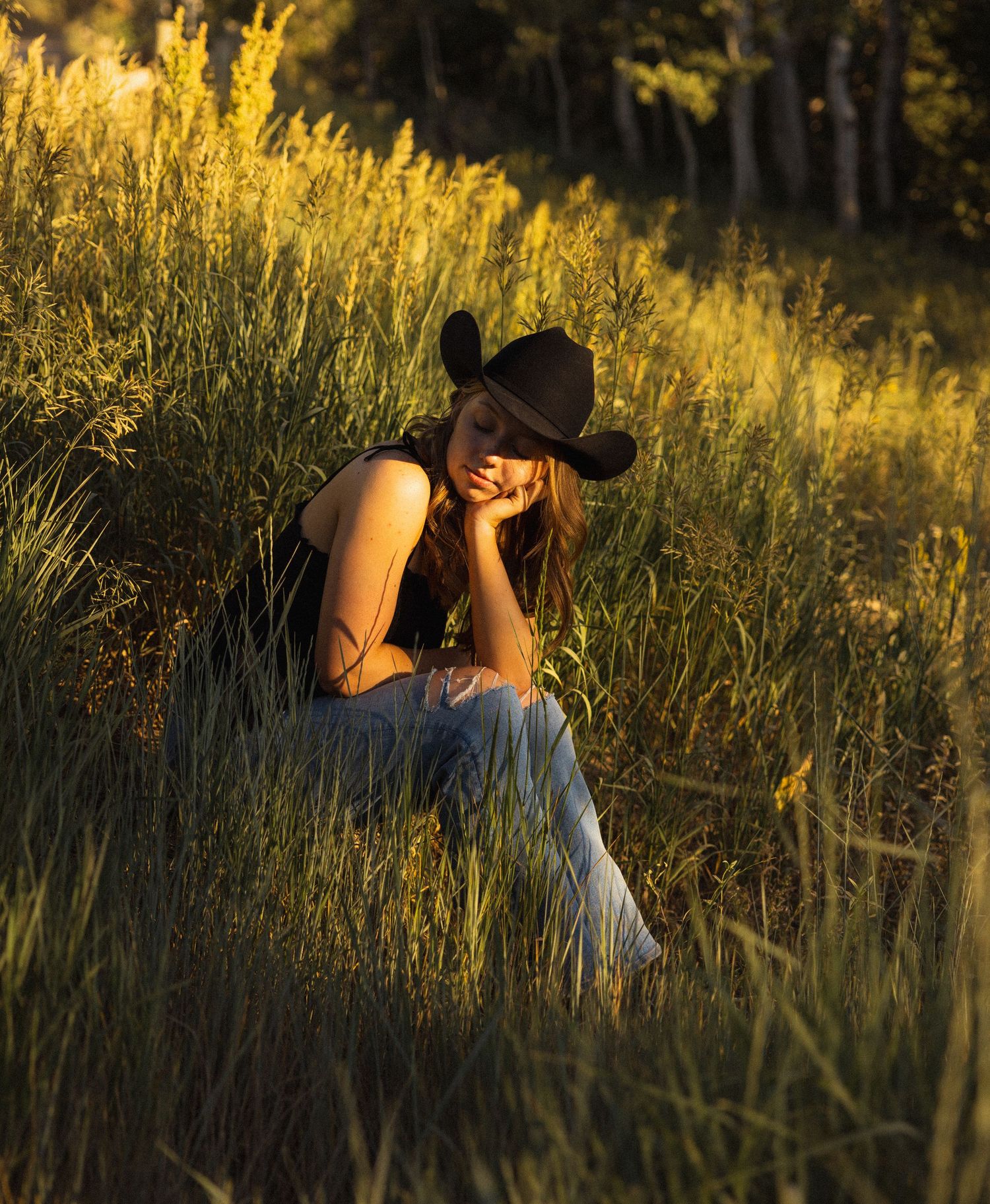 an editorial style photo of a senior girl wearing a cowboy hat and boots sitting in the grass in the mountains at golden hour