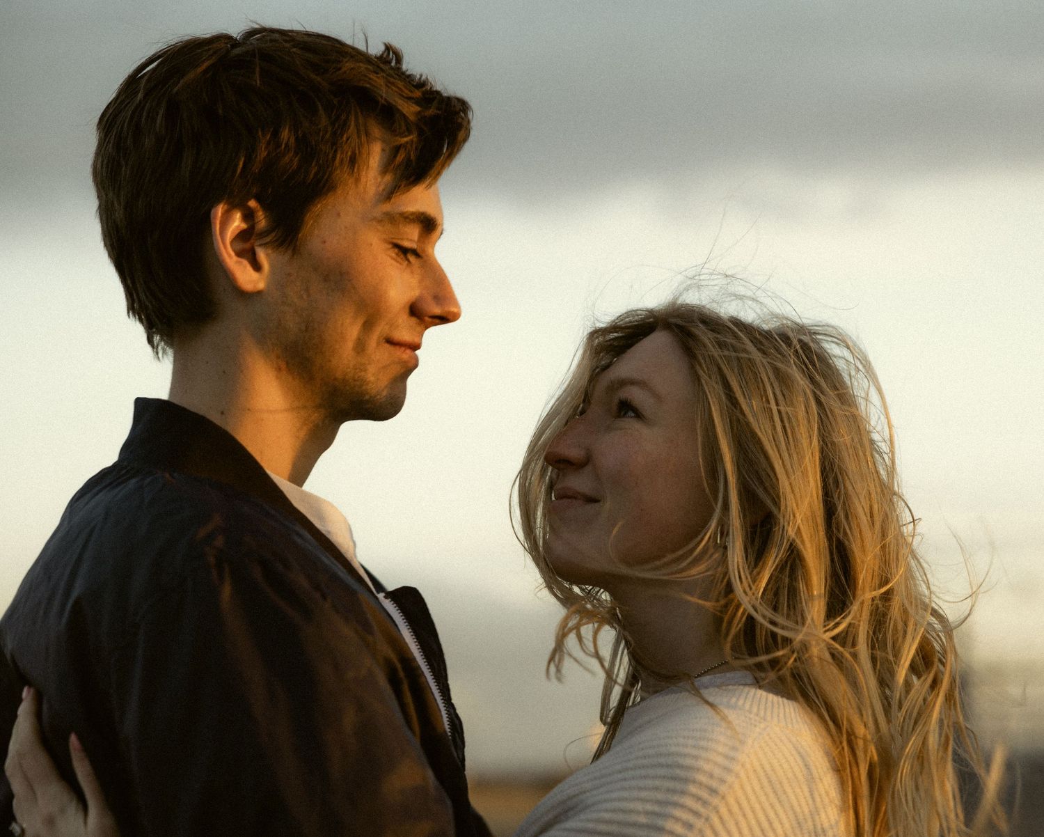 a cinematic filmy photo of a couple looking at each other as the wind blows their hair in their faces with the sun set