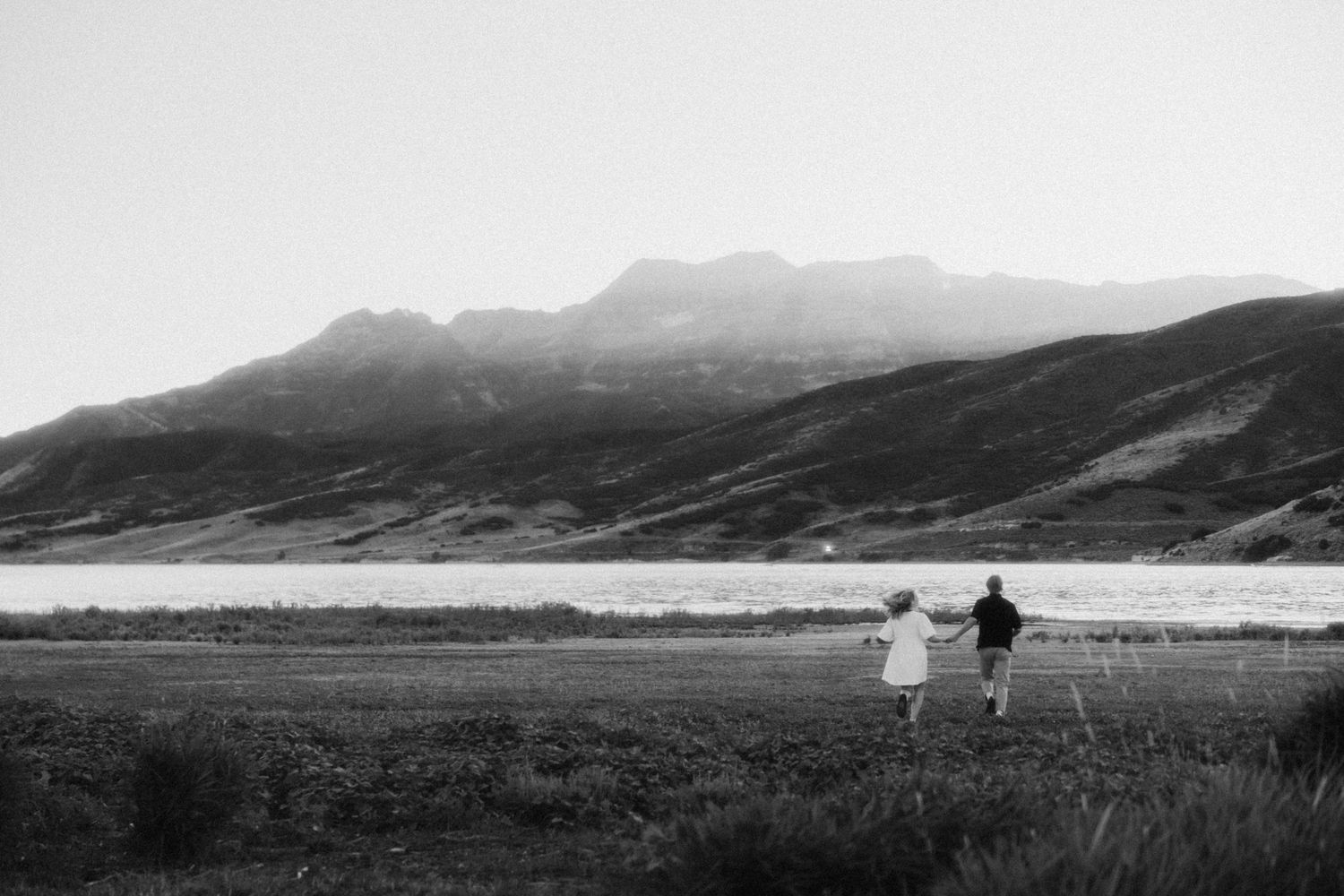 a cinematic black and white photo of a couple running off together with mountains all around and near a lake