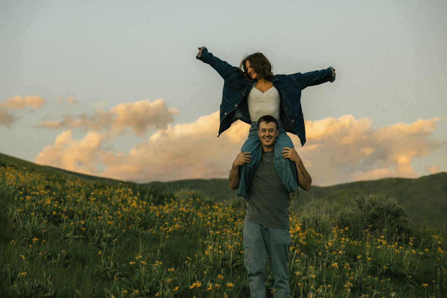 a cinematic filmy photo of a couple running around a wildflower field in the mountains with large fluffy sun lit clouds looming over them