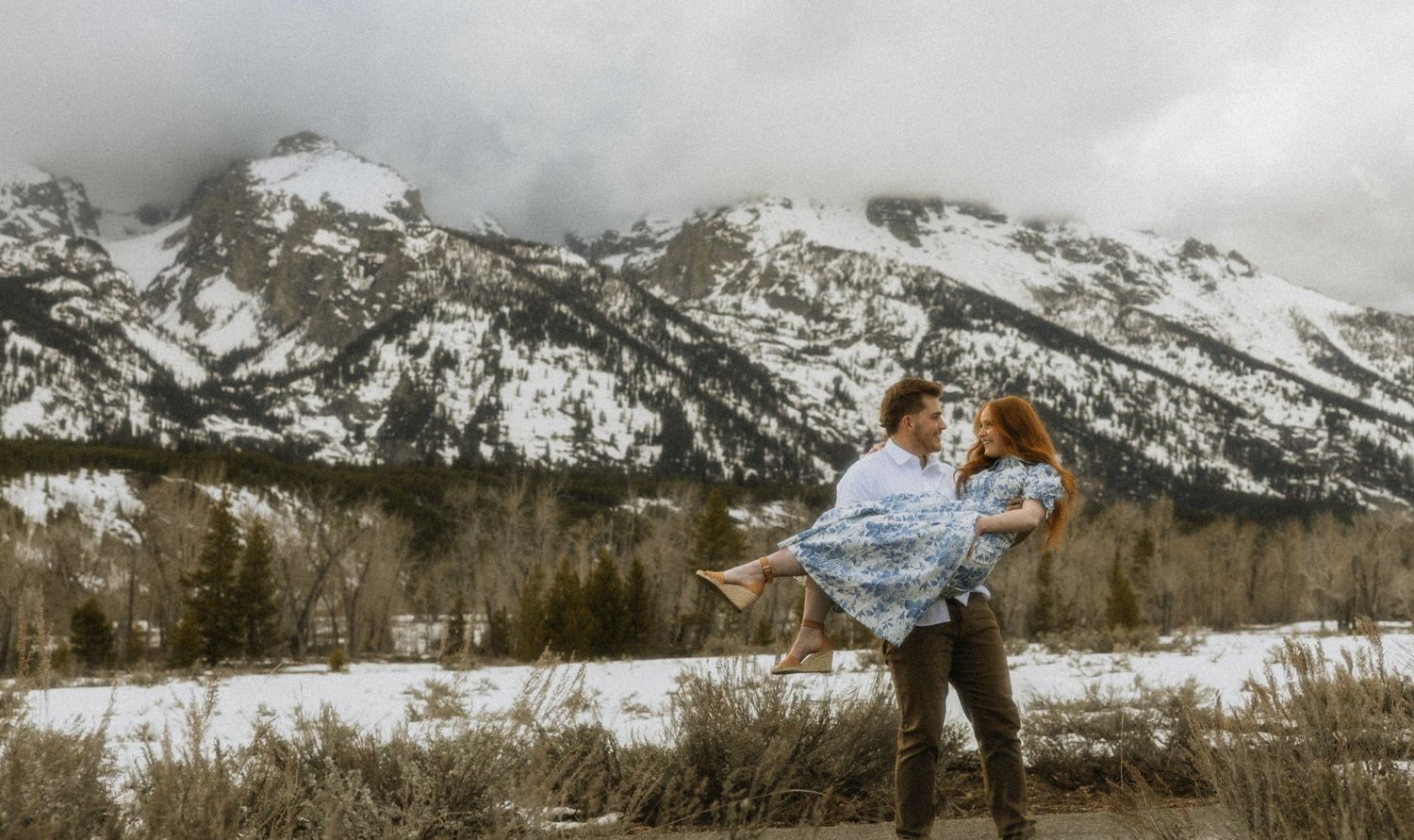 a cinematic filmy photo of a man holding his wife in front of the Teton mountains