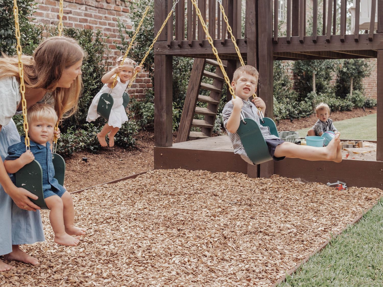 Children play and swing at a rustic outdoor playground with wood chips and brick wall backdrop.