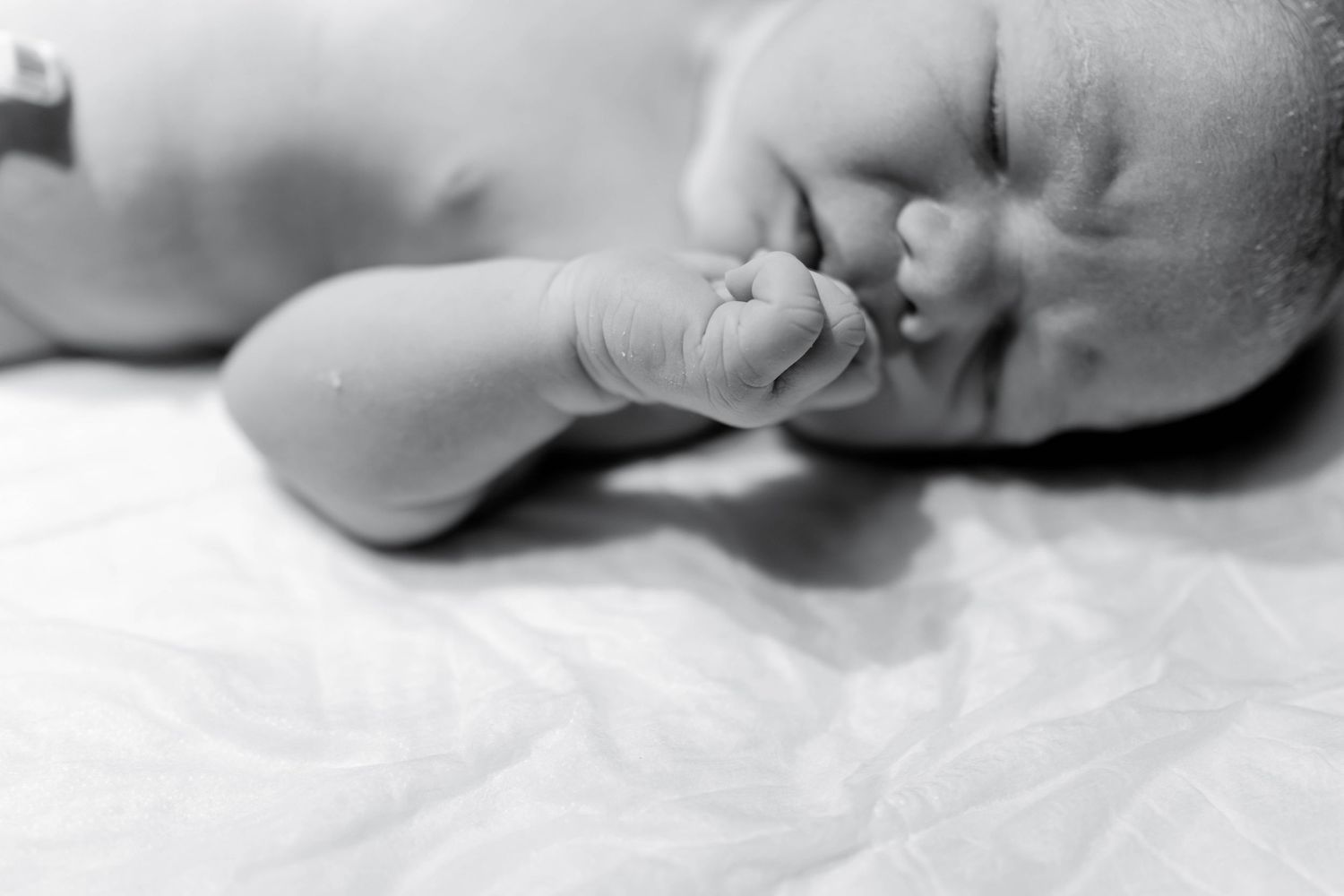 Black and white newborn baby sleeping peacefully, tiny fist raised near face, lying on soft white fabric.