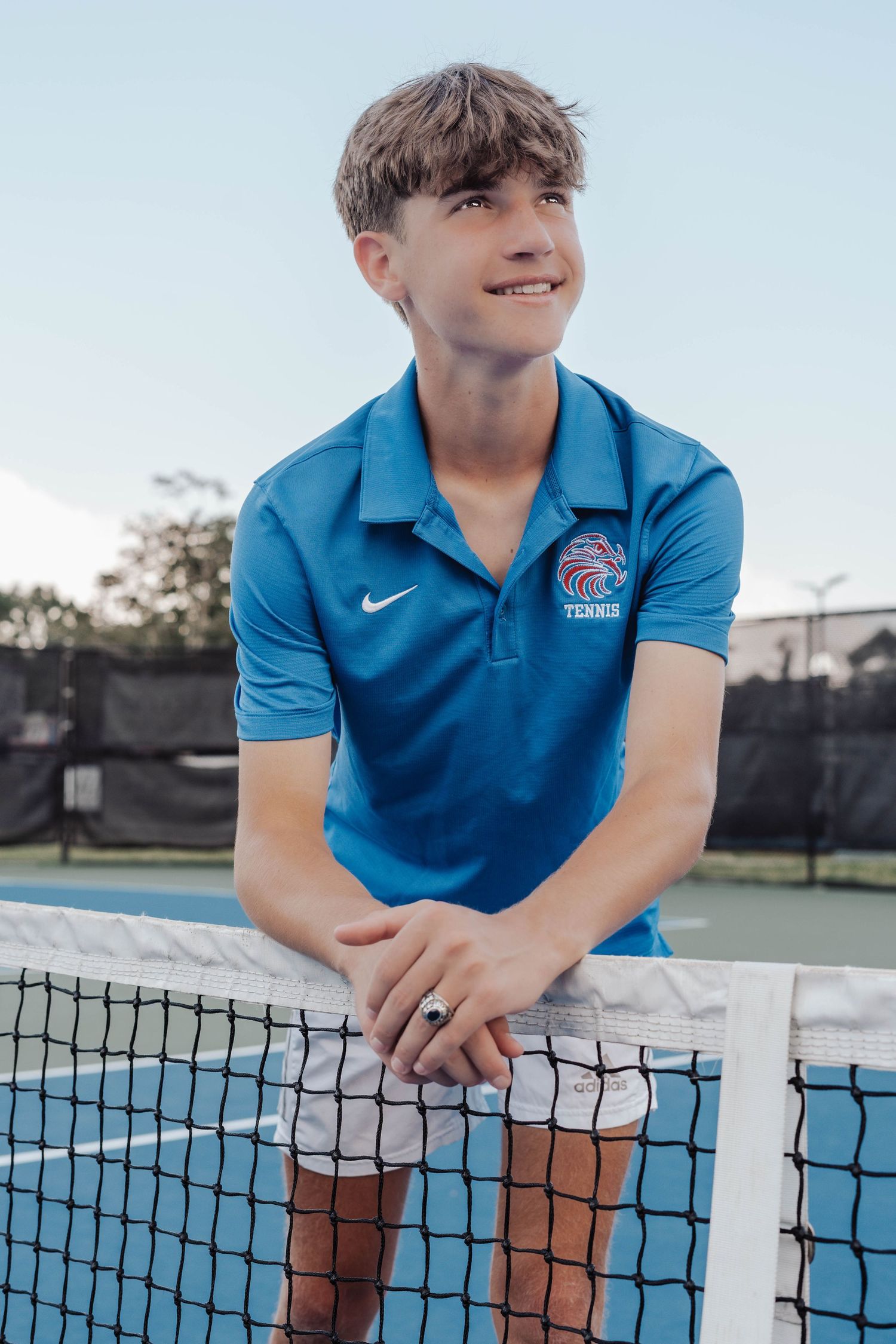 Young male tennis player in blue Nike polo leans on court net, smiling confidently outdoors.