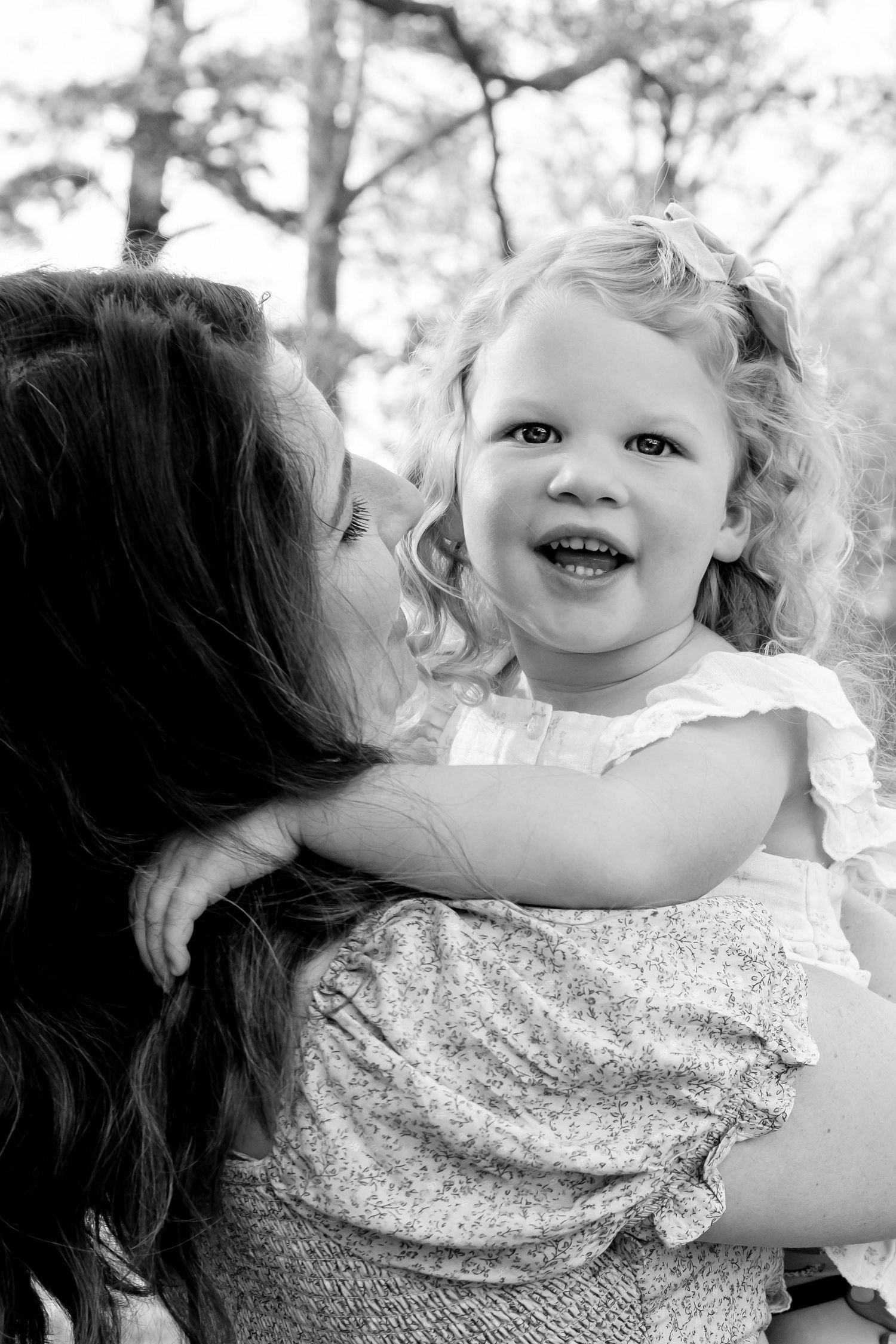 Black and white photo of a mother kissing her smiling toddler girl with curly hair outdoors.