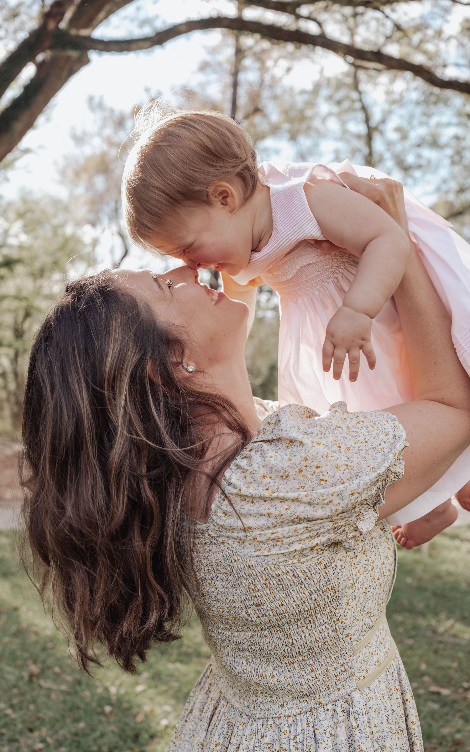 A mother lifts her baby girl in a pink dress outdoors, sharing a sweet nose-to-nose moment.