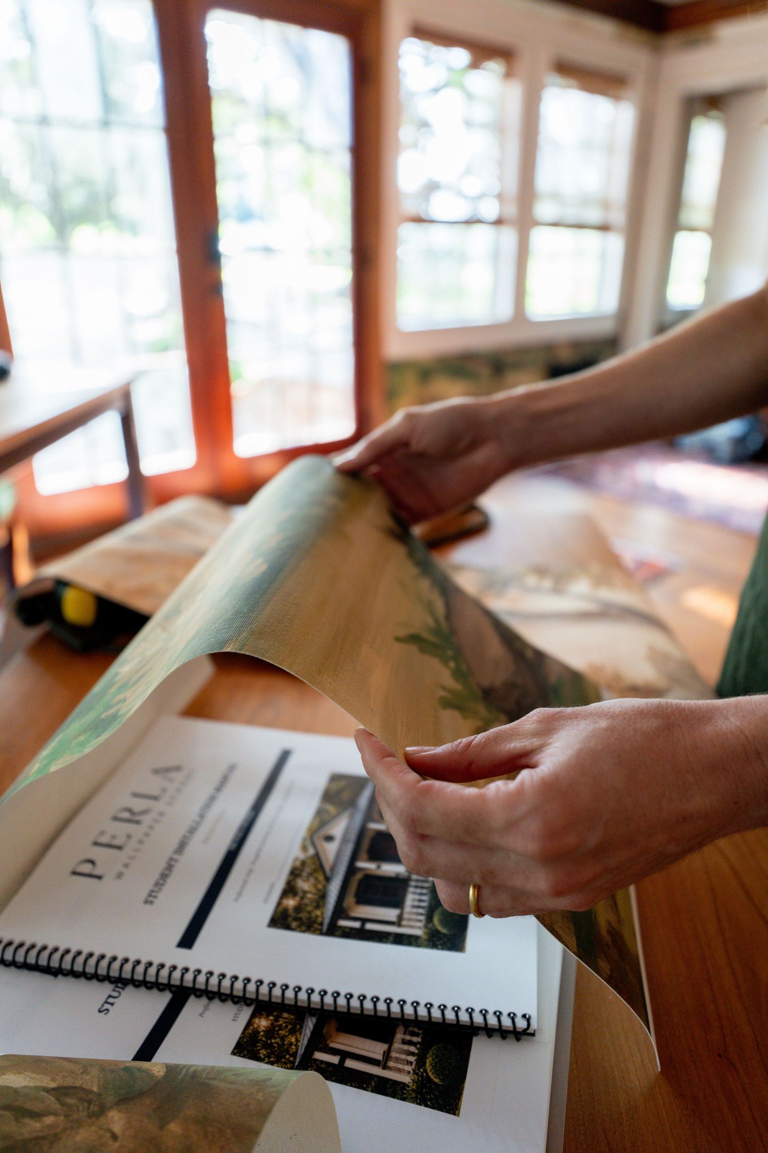 Hands unroll a large printed map beside a spiral-bound Perla brochure on a wooden table in a sunlit room with large windows.