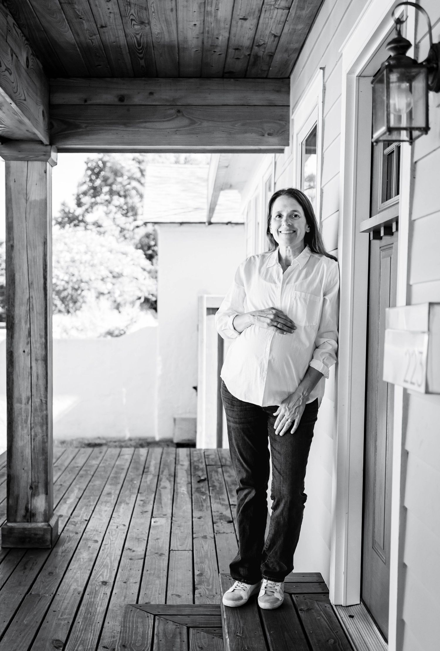 Black and white photo of a smiling pregnant woman in white shirt and dark pants standing on a wooden porch.