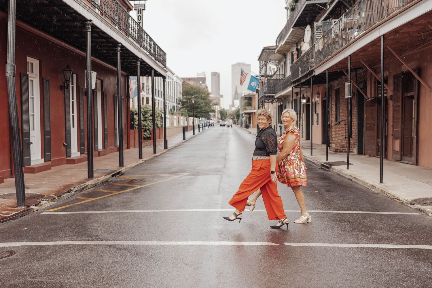 Two women crossing a wet street in New Orleans French Quarter with historic buildings lining both sides.
