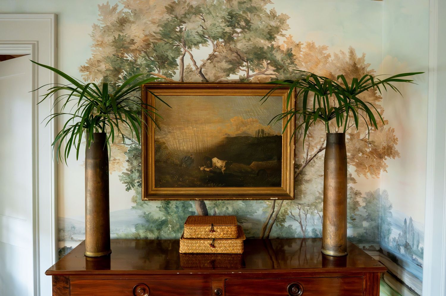Antique wooden console table with a framed landscape painting, tall bronze vases holding palm fronds, and wicker boxes against a hand-painte