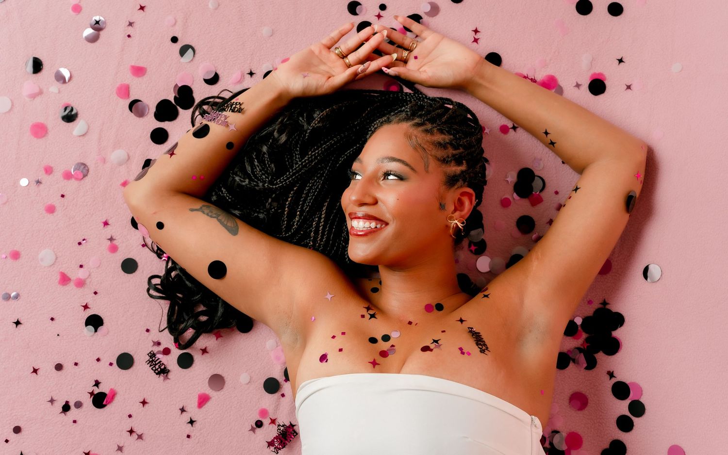 Young Black woman smiling joyfully lying on pink background with colorful confetti falling around her.