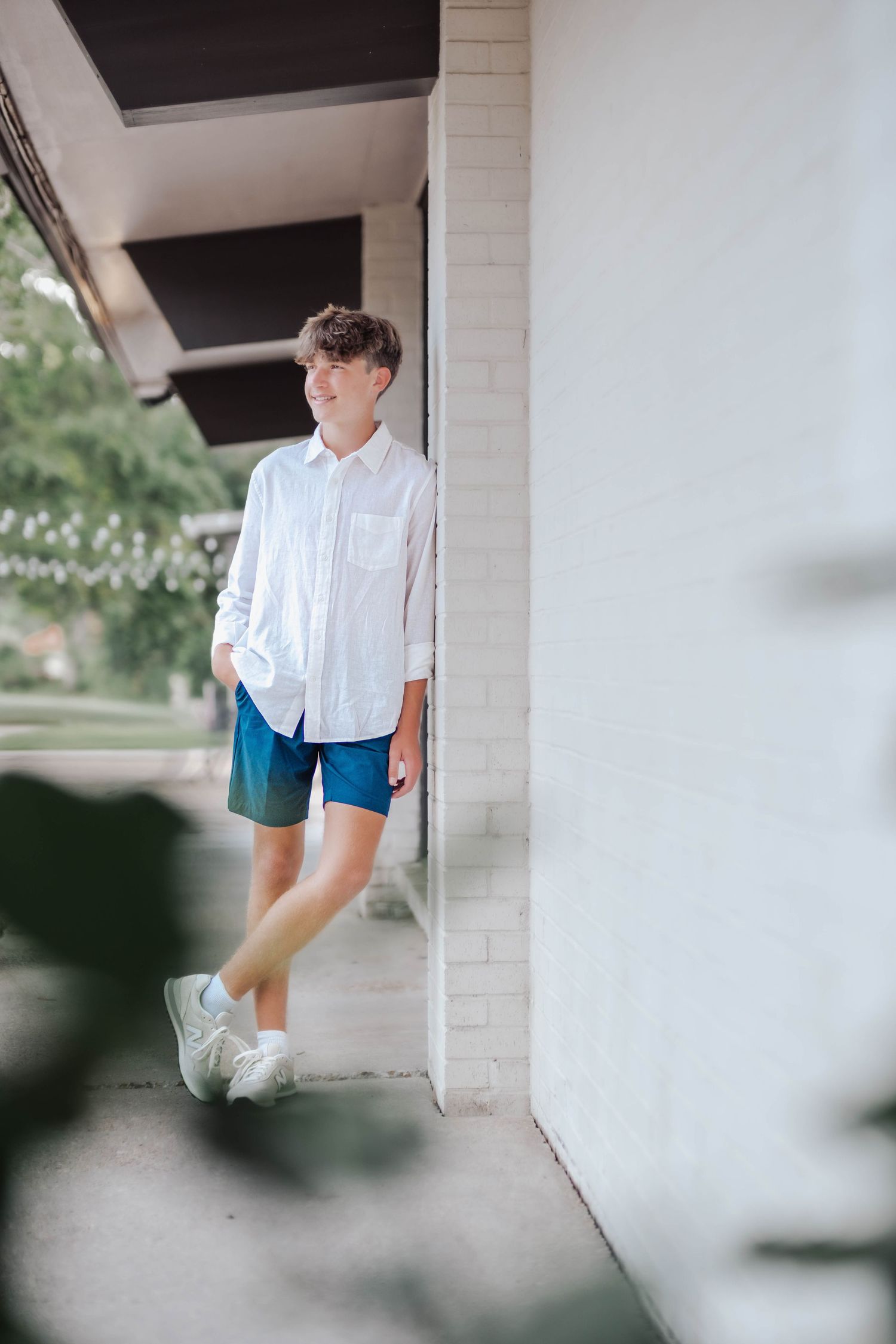 A teenage boy in a white shirt and blue shorts leans against a white brick wall outdoors.