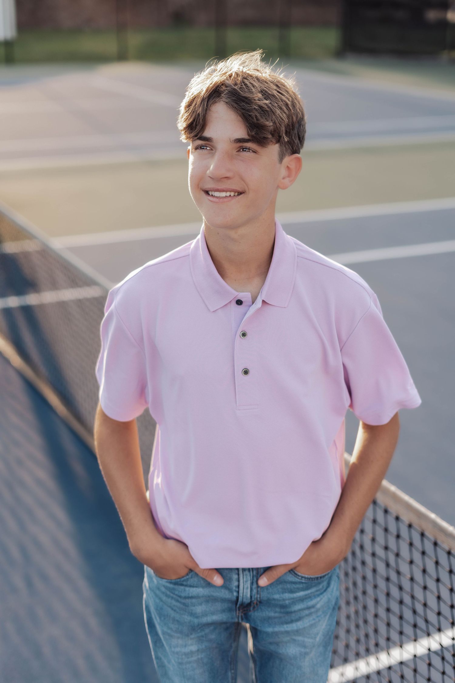 Smiling teenage boy in pink polo shirt and jeans standing on a tennis court near the net on a sunny day.