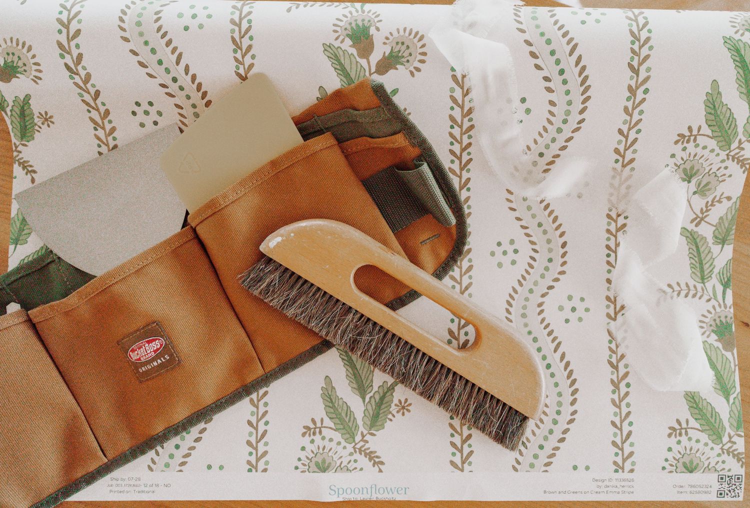 Canvas tool pouch with a wooden-handled wallpaper smoothing brush and scrapers resting on a green floral striped wallpaper sheet.