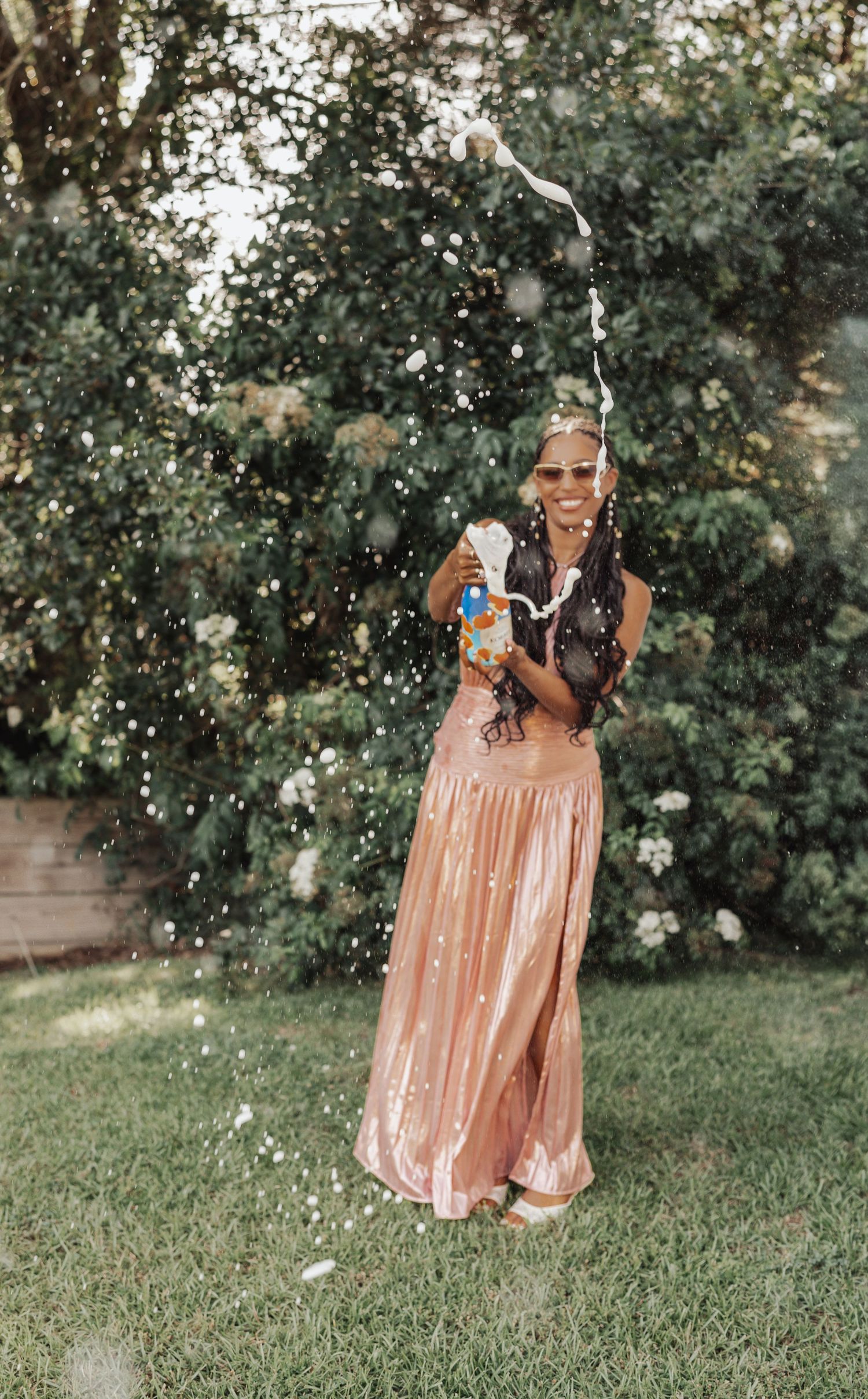 Woman in rose gold skirt spraying water bottle in lush green backyard, wearing sunglasses and braids.