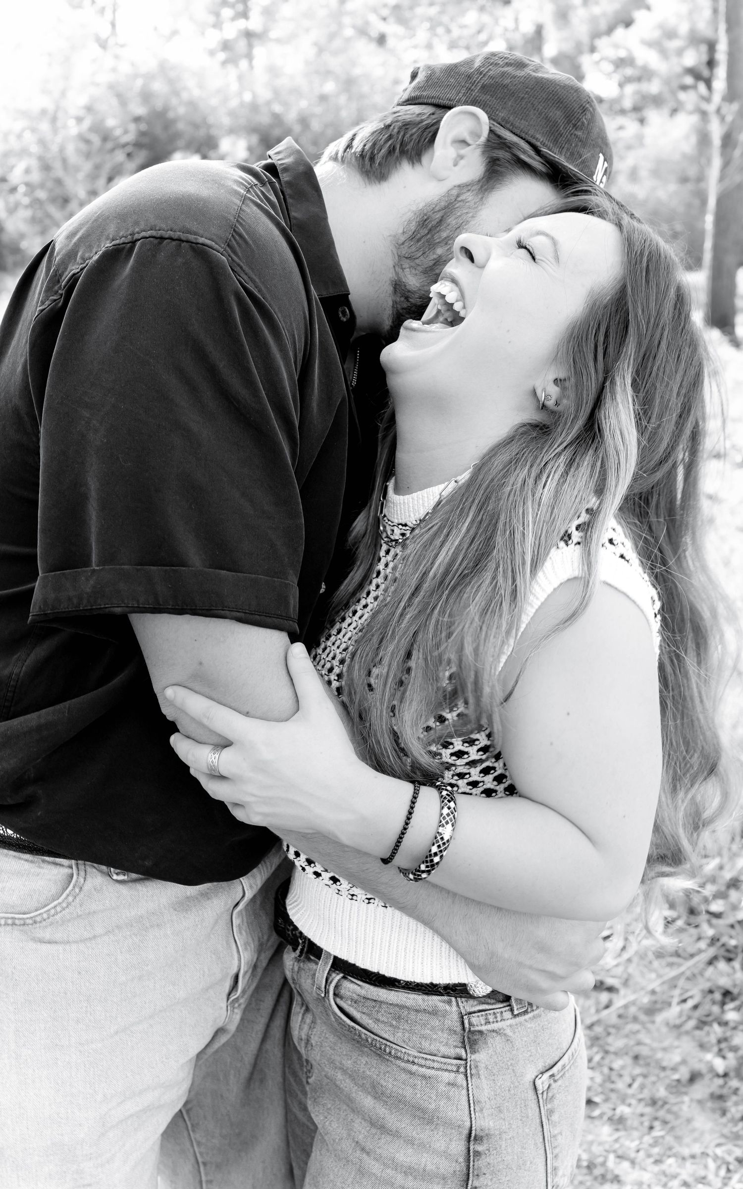 Black and white photo of a laughing couple embracing outdoors, woman in crochet top and jeans, man in dark shirt and cap.
