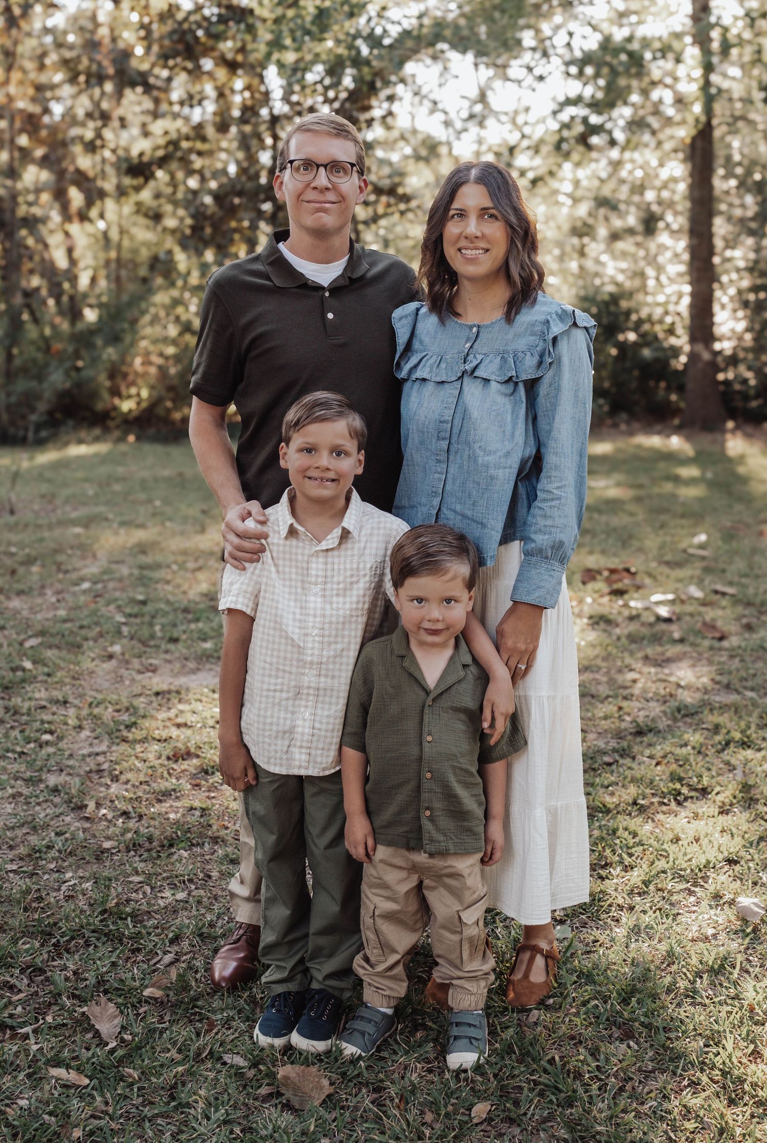 Family of four posing outdoors in a park, parents standing behind two young boys on green grass.