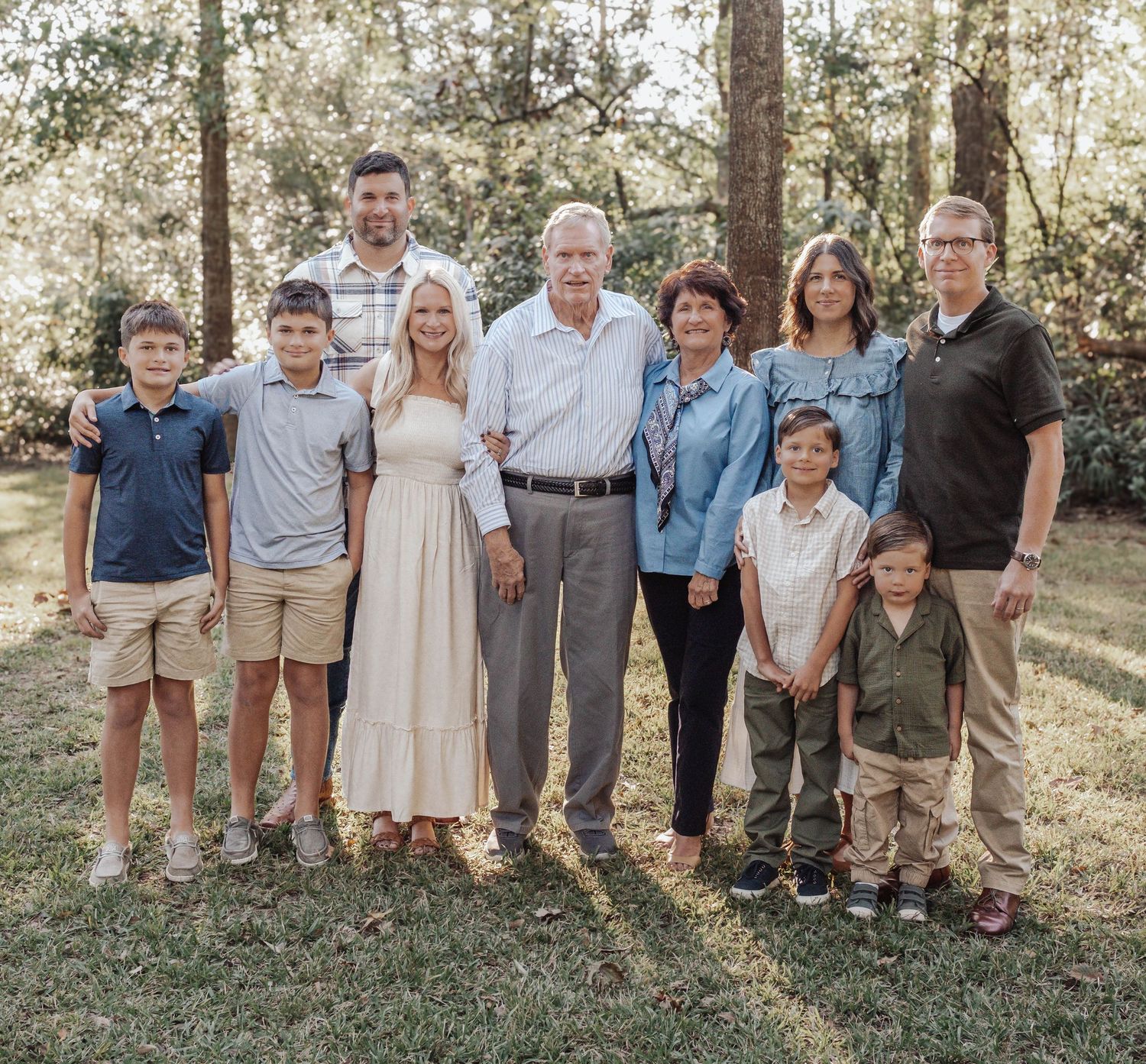 A multigenerational family of nine poses together outdoors on grass surrounded by lush green trees.