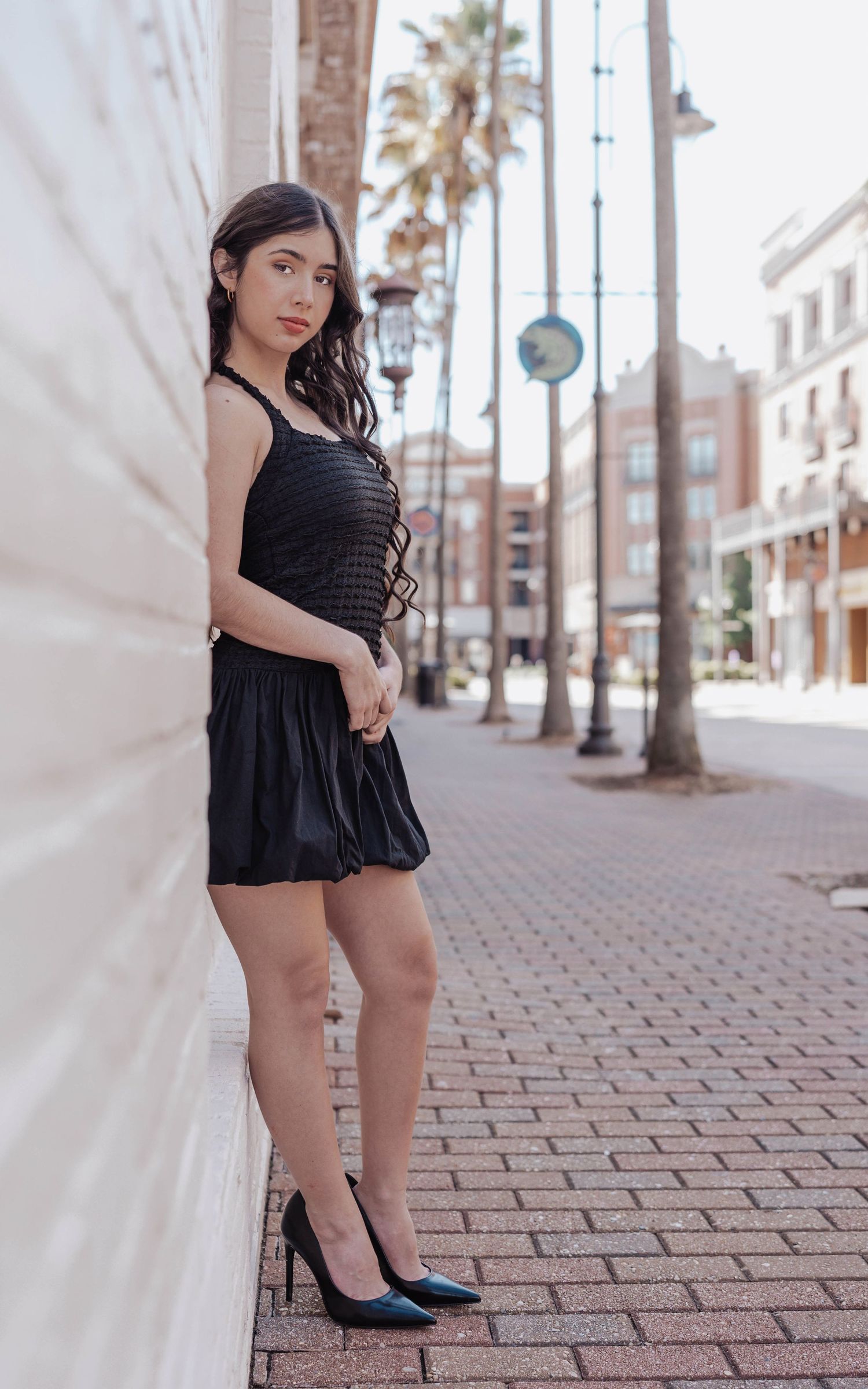 Young woman in black mini dress and heels leaning against white wall on brick sidewalk with palm trees.