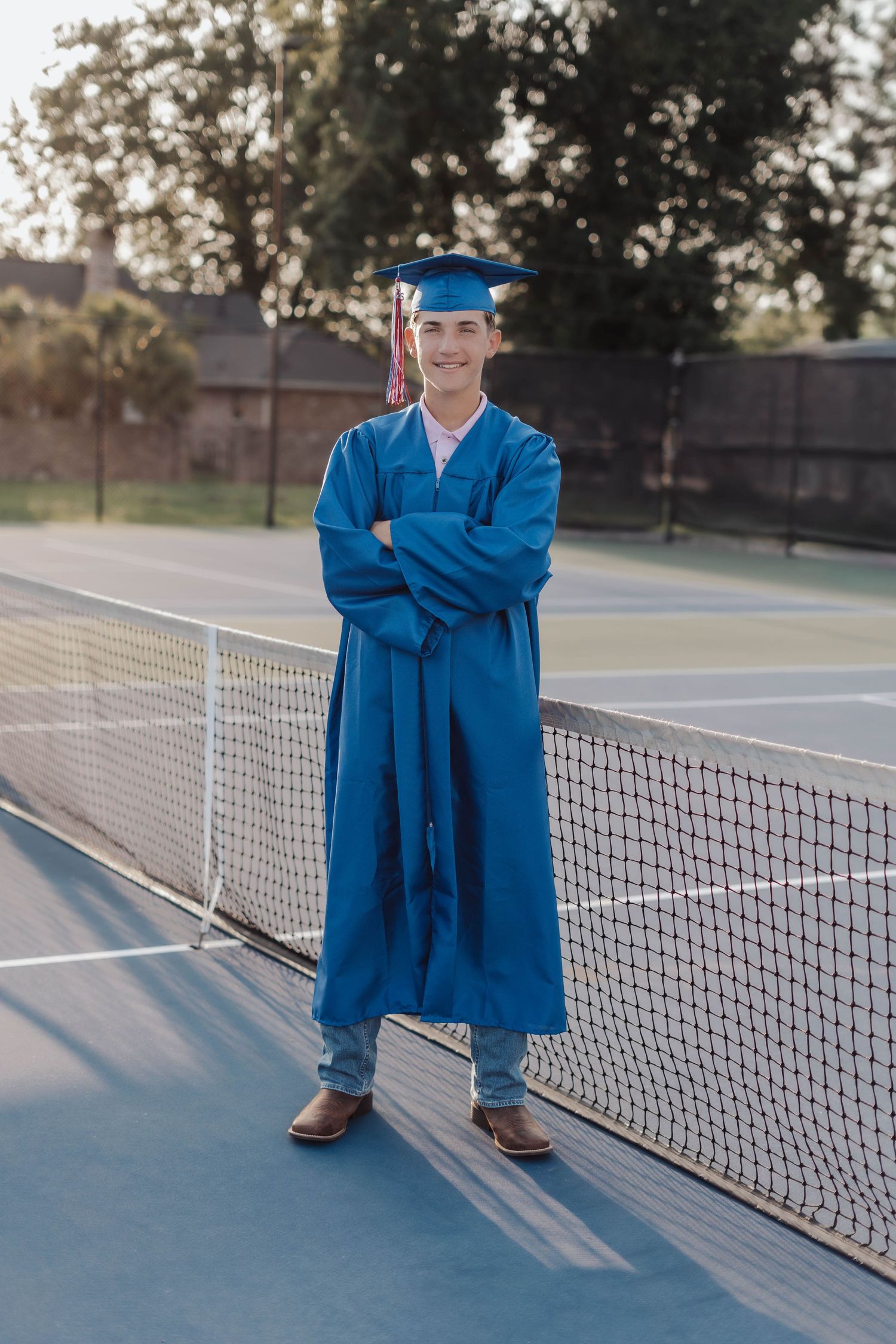 Young graduate in blue cap and gown stands confidently on tennis court with arms crossed, wearing cowboy boots.