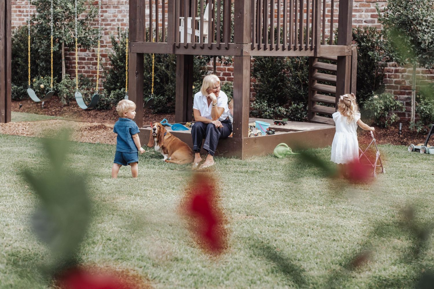 Children playing in a backyard with a wooden playset on green grass near a brick house.