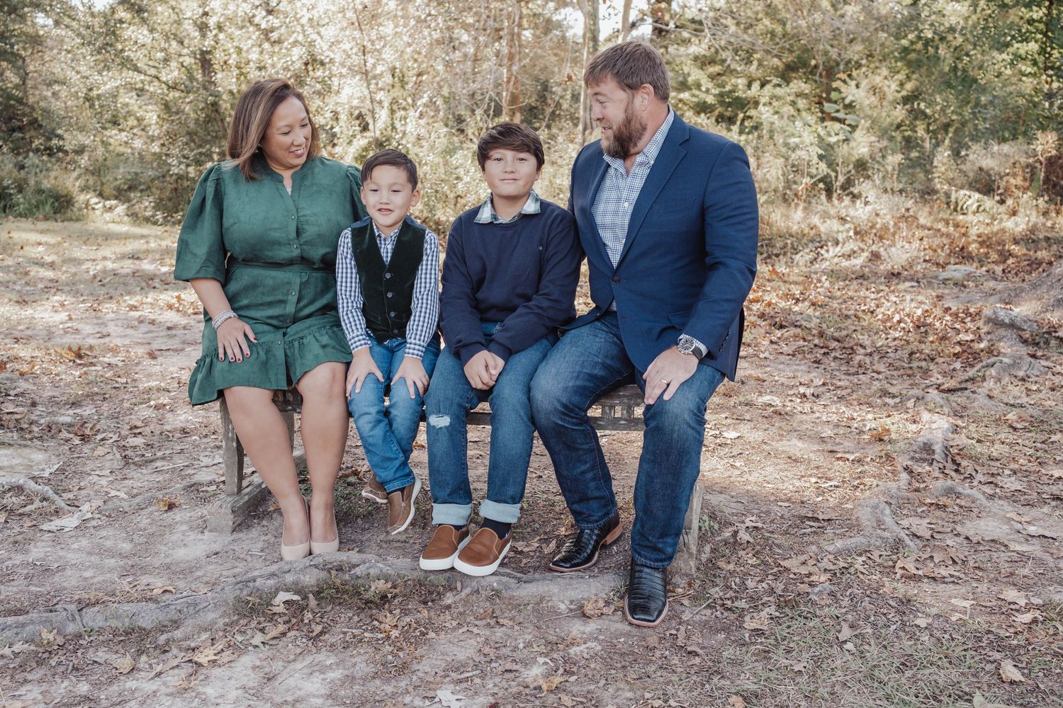 Family of four sitting on a log in a wooded outdoor setting, smiling and enjoying a candid moment together.