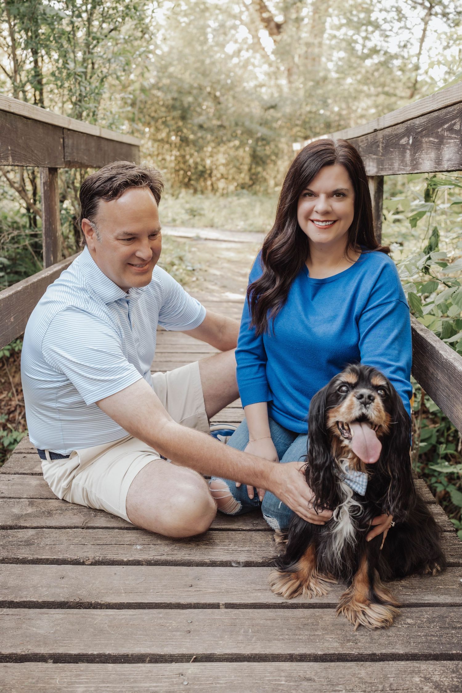 Couple sitting on wooden bridge with Cavalier King Charles Spaniel dog in a lush green outdoor setting.