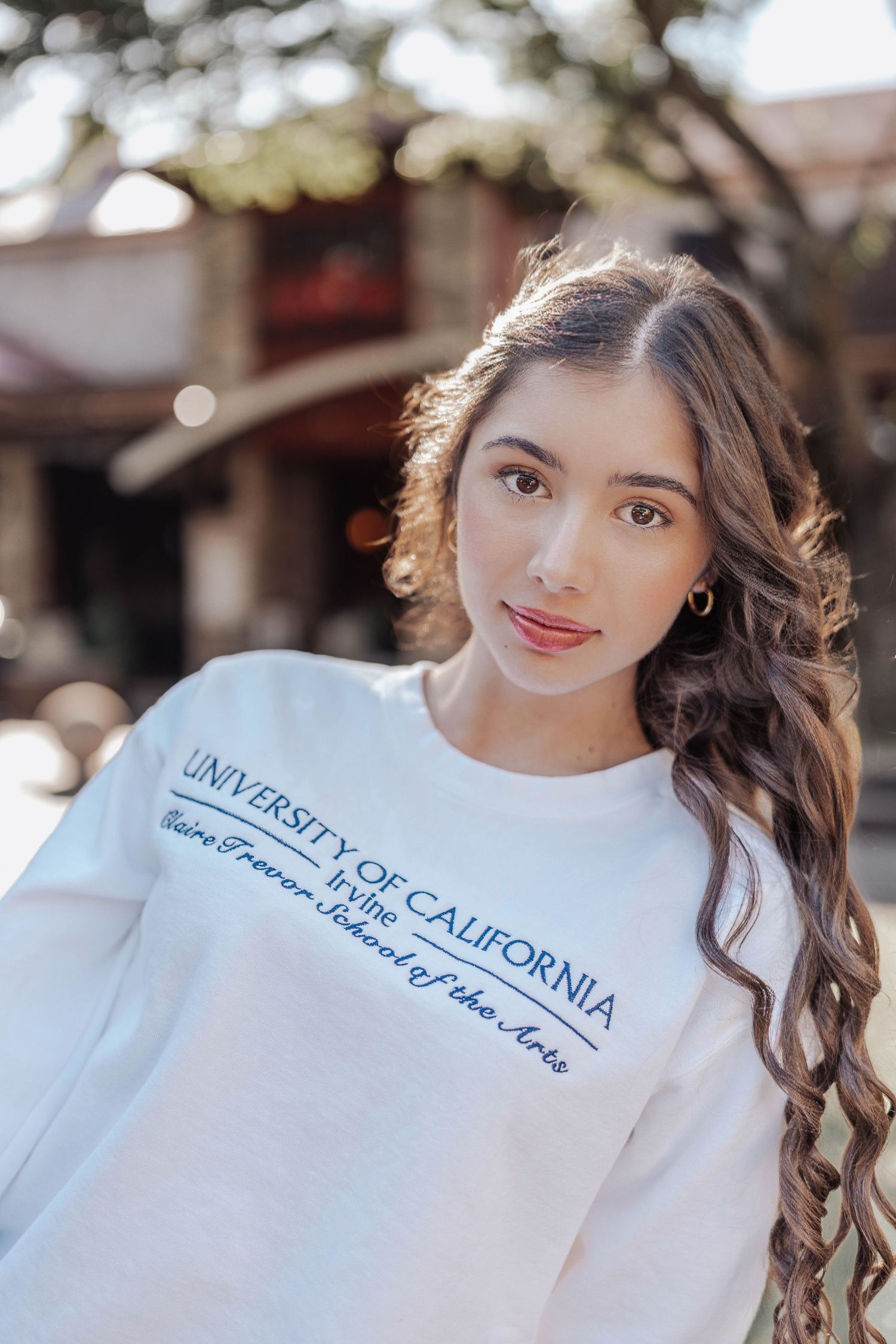 Young woman wearing a white University of California Irvine Claire Trevor School of the Arts sweatshirt outdoors.
