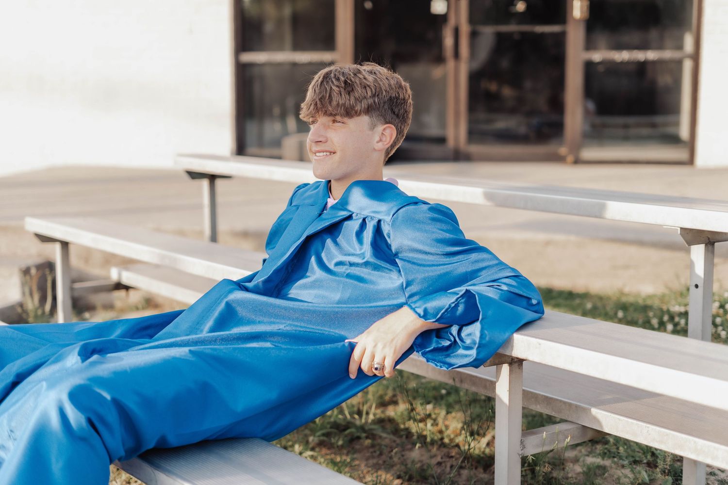 A smiling young boy relaxes on metal bleachers wearing an oversized blue satin graduation gown outdoors.