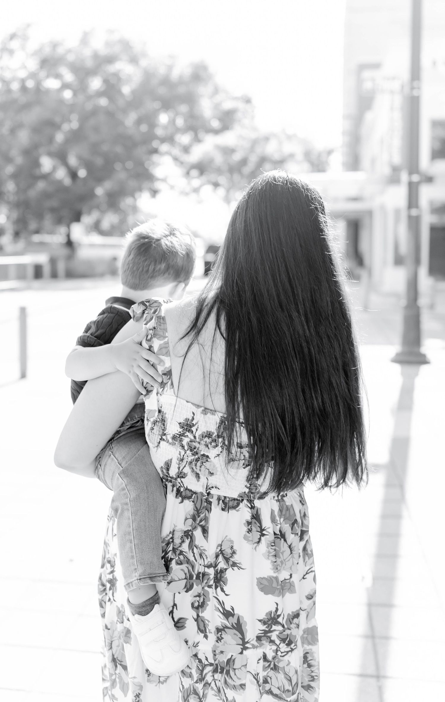 Black and white photo of a mother with long dark hair holding her young child outdoors in a floral dress.