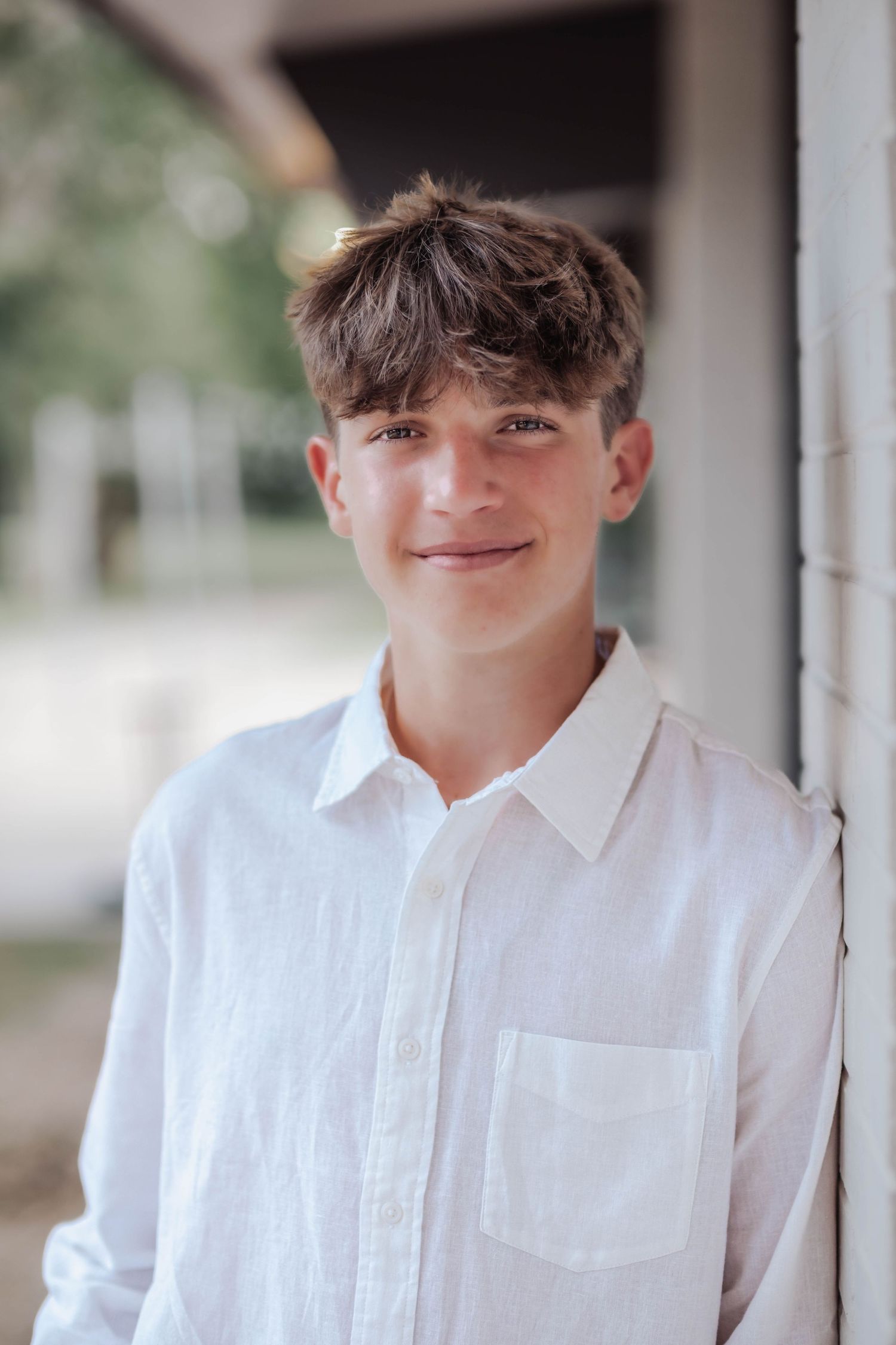A smiling teenage boy in a white linen shirt leans against a white wall outdoors.