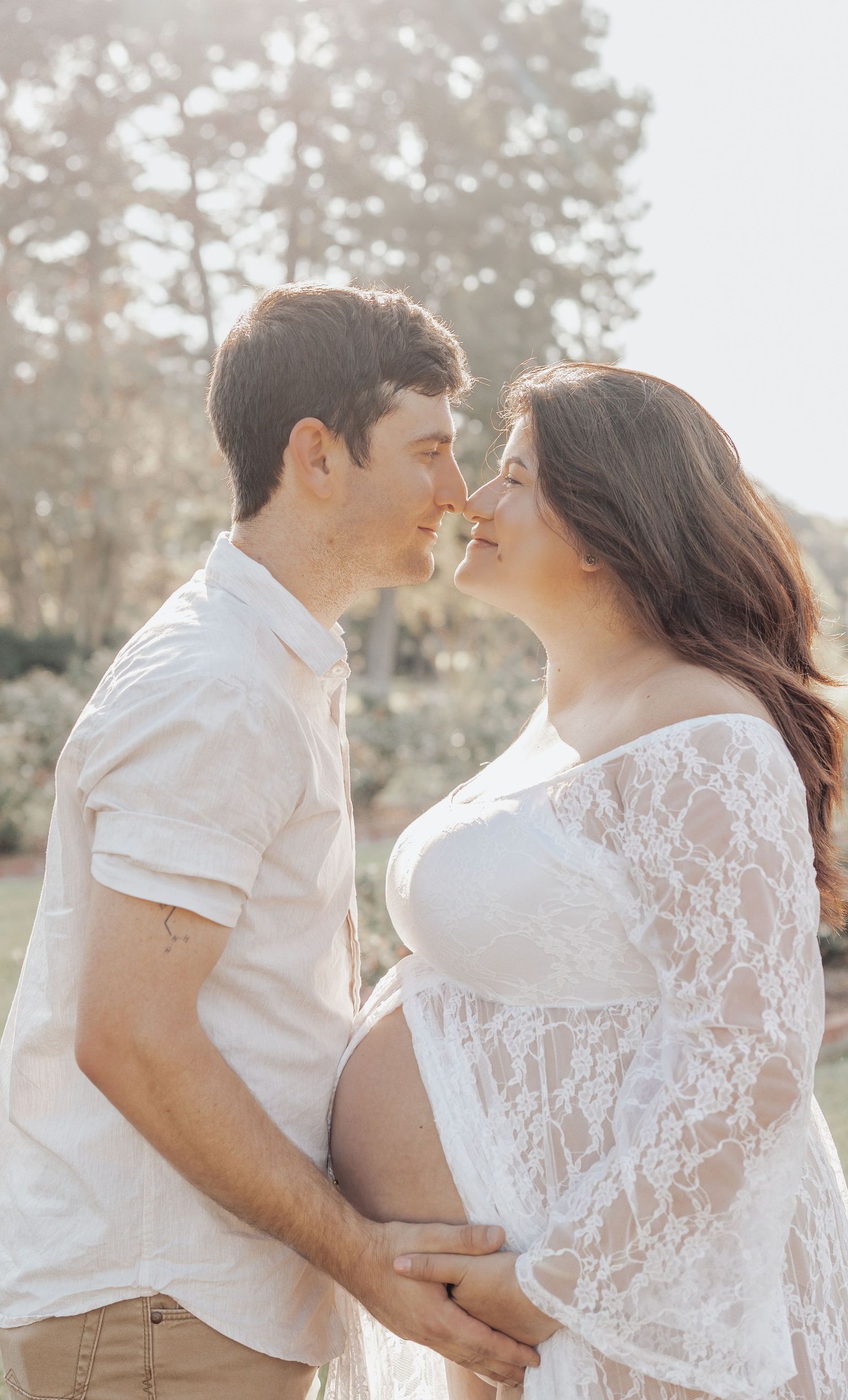 Couple sharing a tender nose-to-nose moment outdoors, woman in lace maternity dress cradling baby bump in soft sunlight.