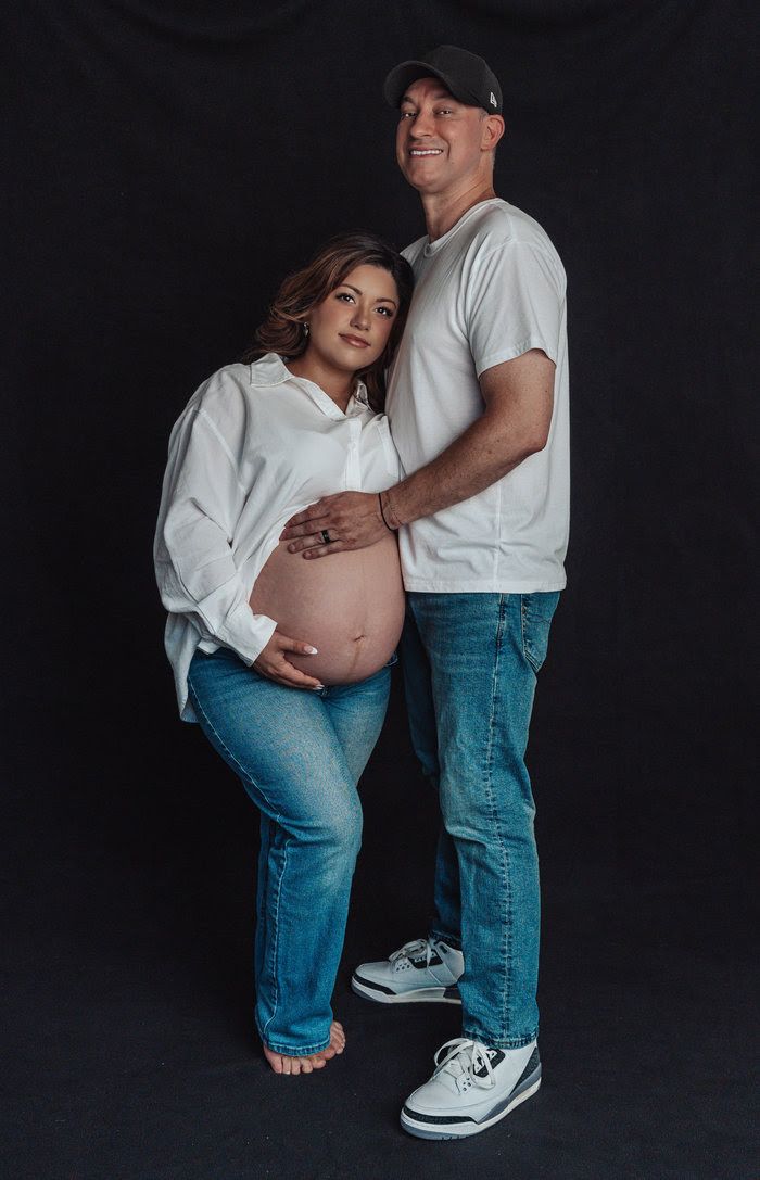 Couple posing for maternity photo shoot against dark background, woman showing baby bump in white shirt and jeans.