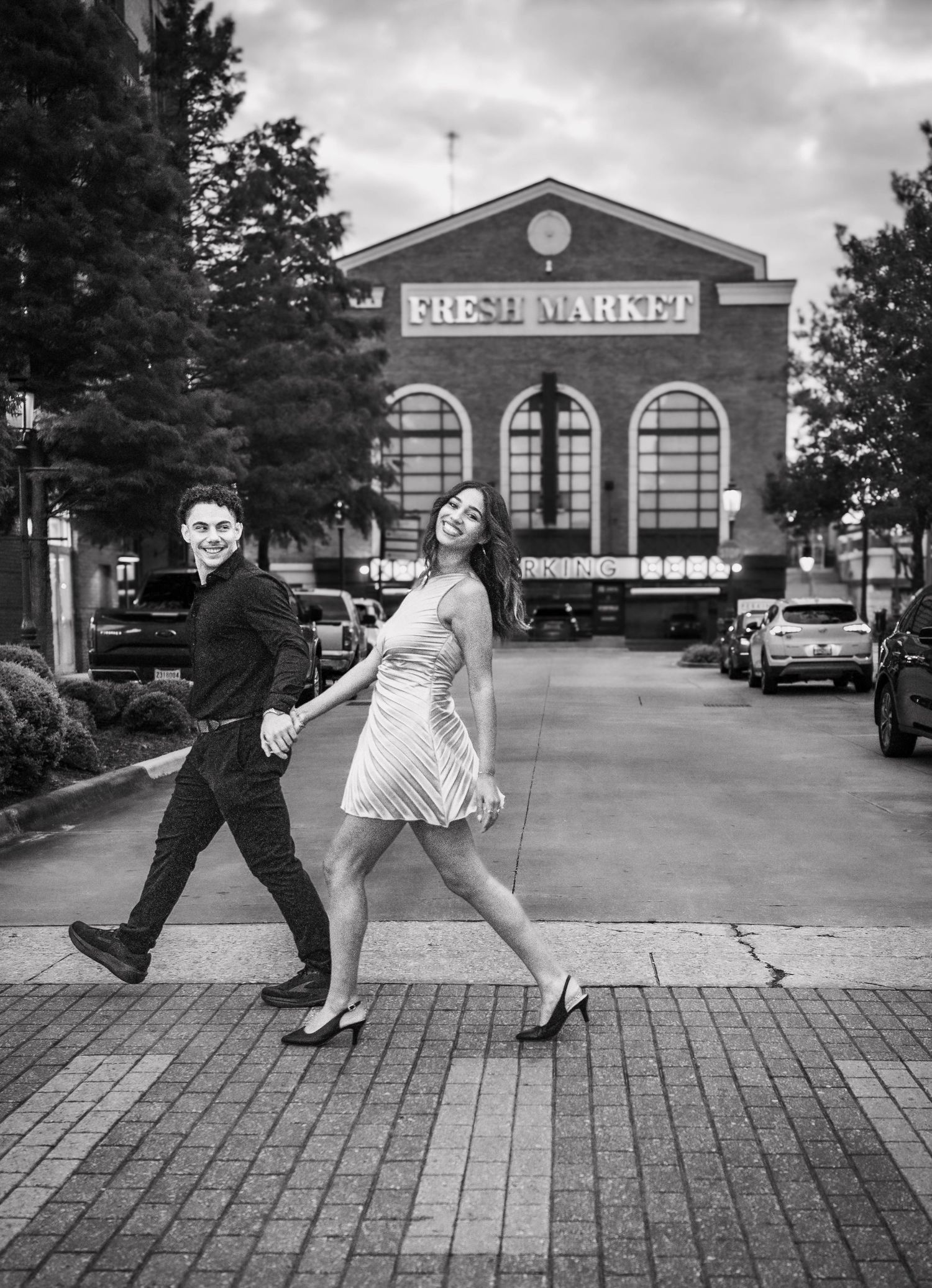 Couple holding hands walking in front of Fresh Market building in black and white engagement photo.
