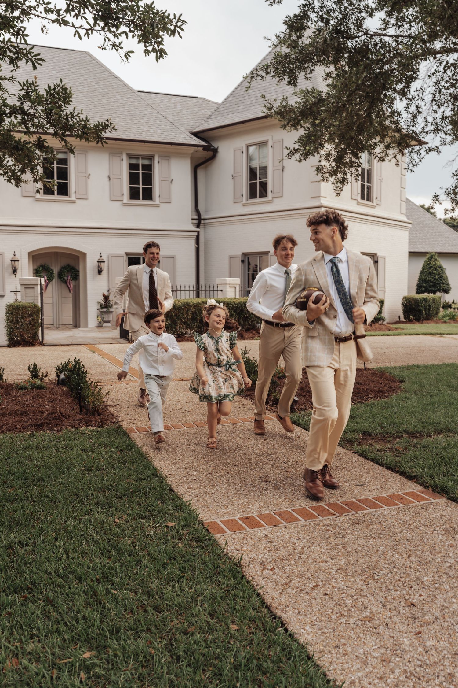 A family in formal attire runs joyfully along a brick path outside a large white estate home.