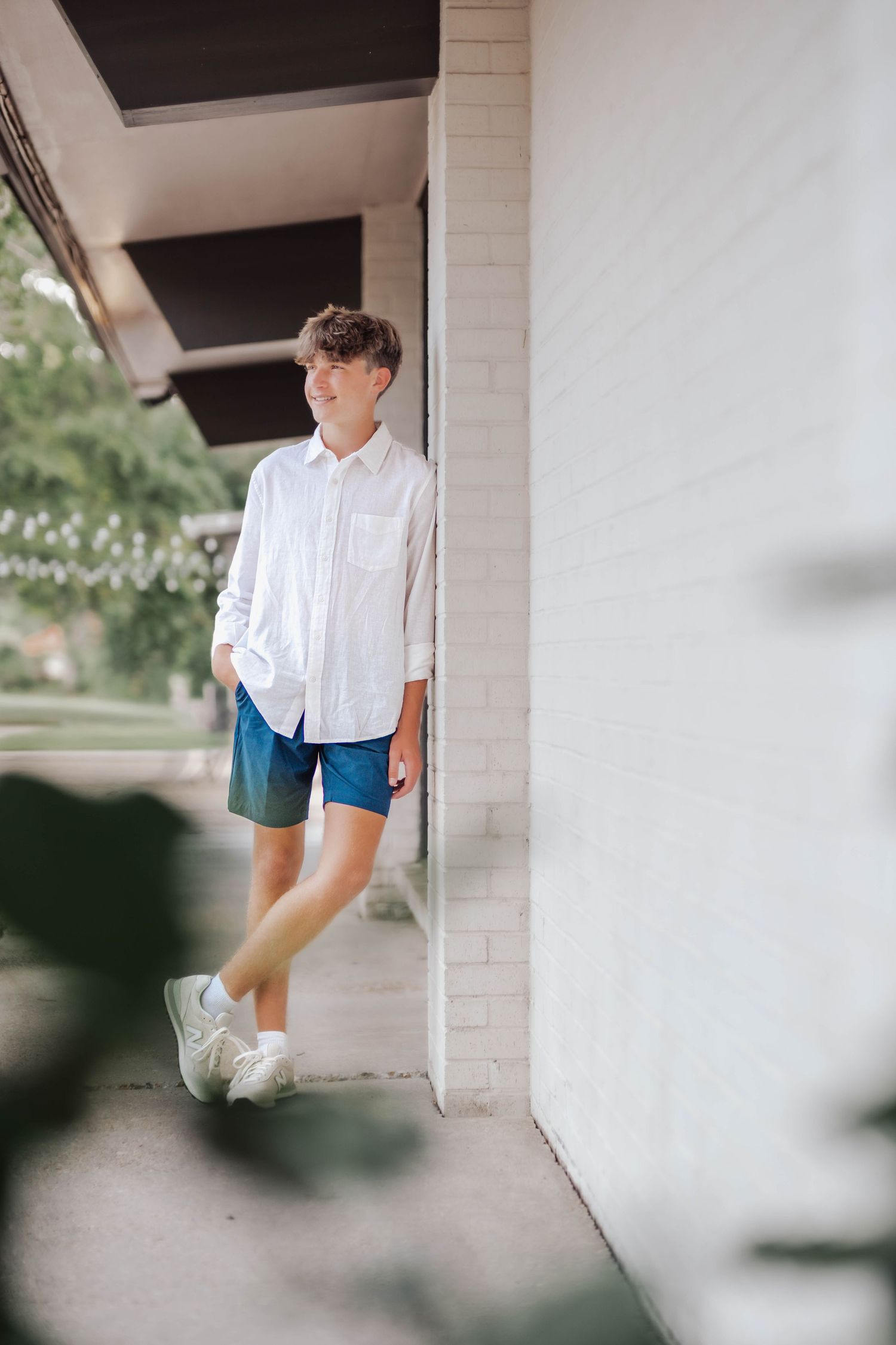 Teen boy in white shirt and blue shorts leaning against white brick wall outdoors, smiling casually.