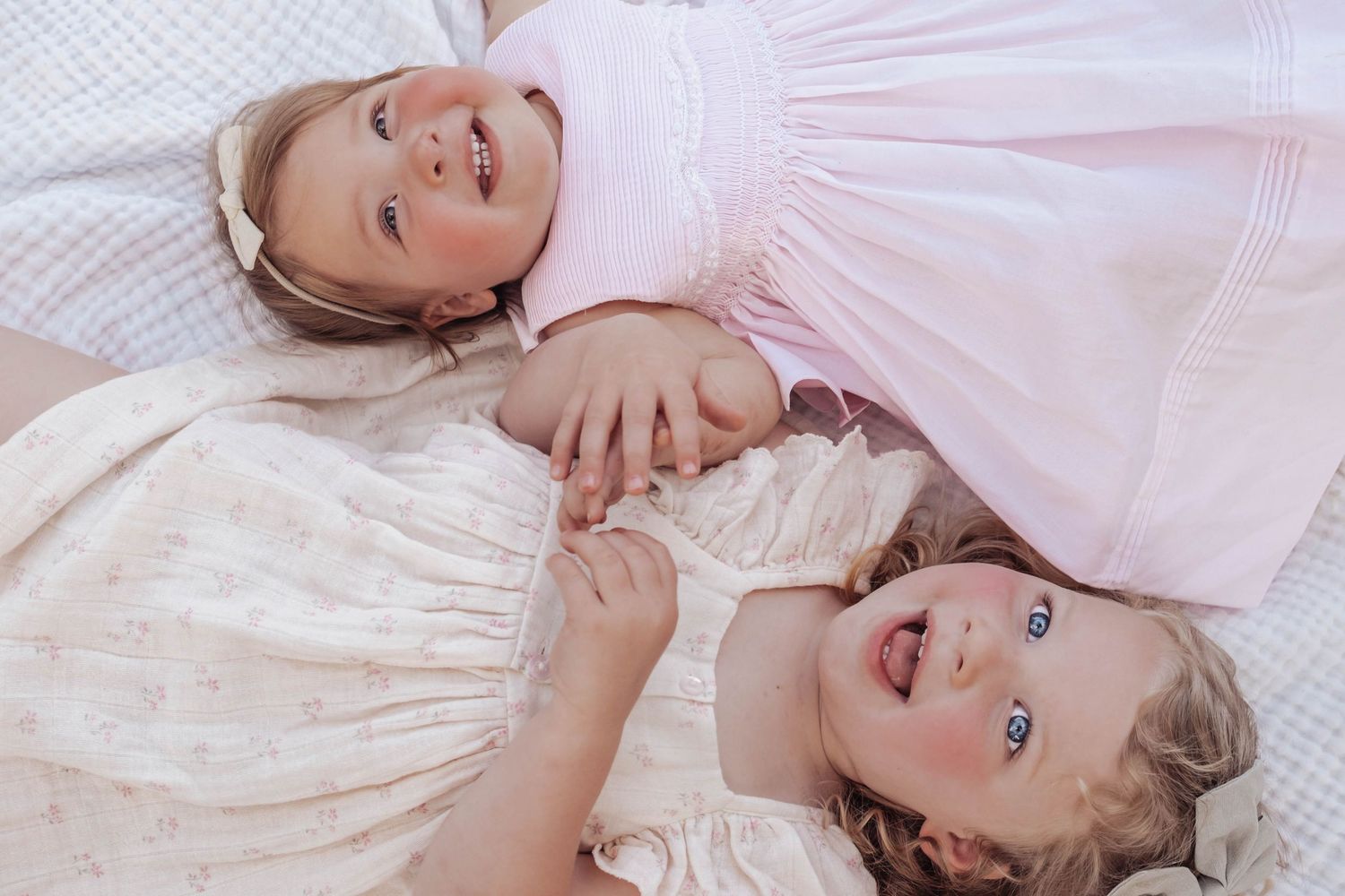 Two smiling baby girls in pink and cream dresses lying together on a white quilted surface.