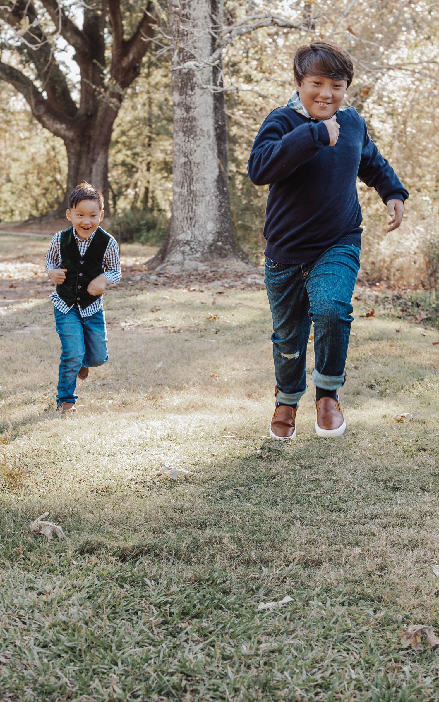 A child in a navy sweater and blue pants jumps playfully on a grassy slope surrounded by trees on an autumn day.