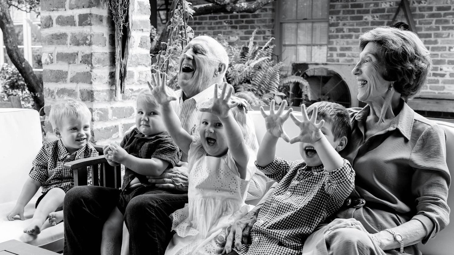Black and white photo of a joyful family group with children making funny faces outdoors near a brick wall.