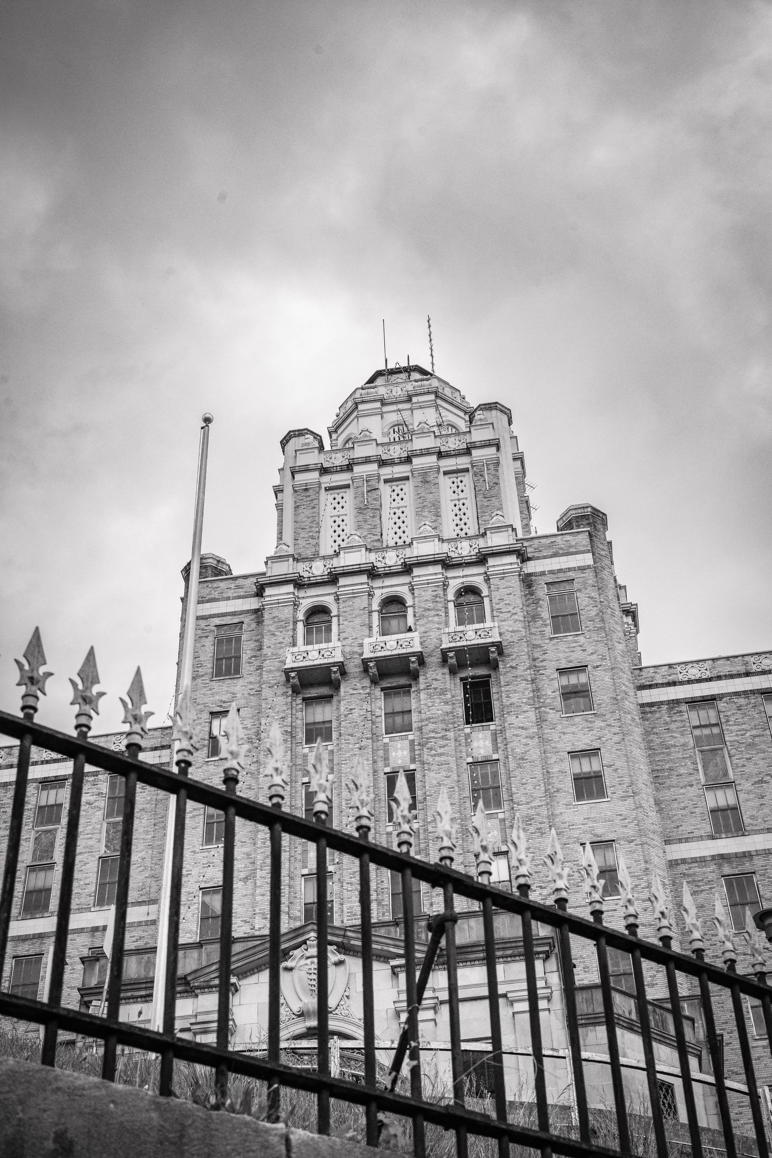 Black and white photo of an ornate historic building with decorative facade and iron fence in foreground under cloudy sky.