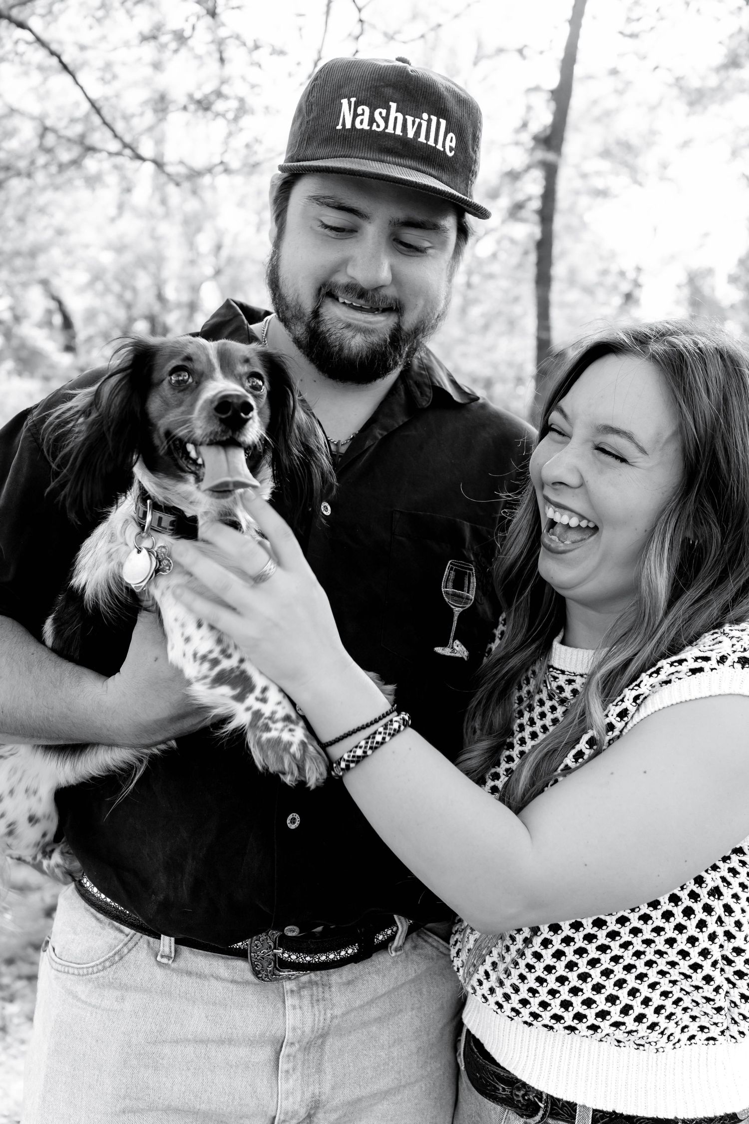 Black and white photo of smiling couple holding a spaniel dog outdoors, man wearing Nashville cap, woman laughing joyfully.