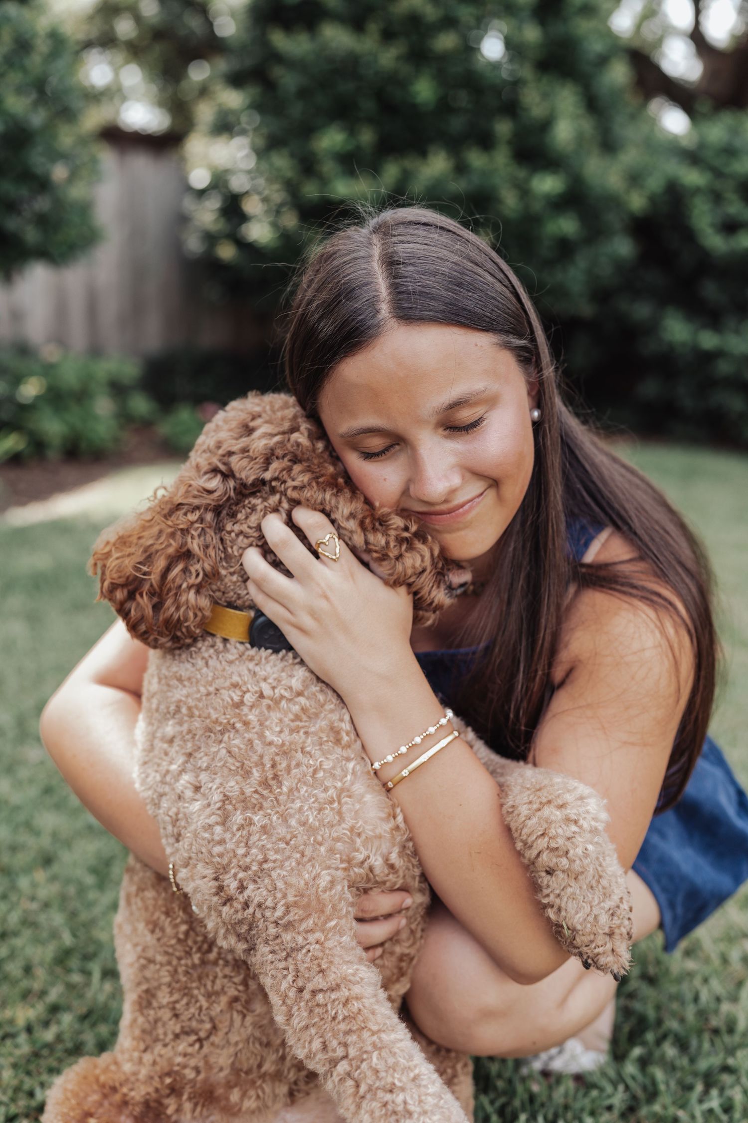 A smiling teenage girl hugs her fluffy golden doodle puppy in a lush green backyard.