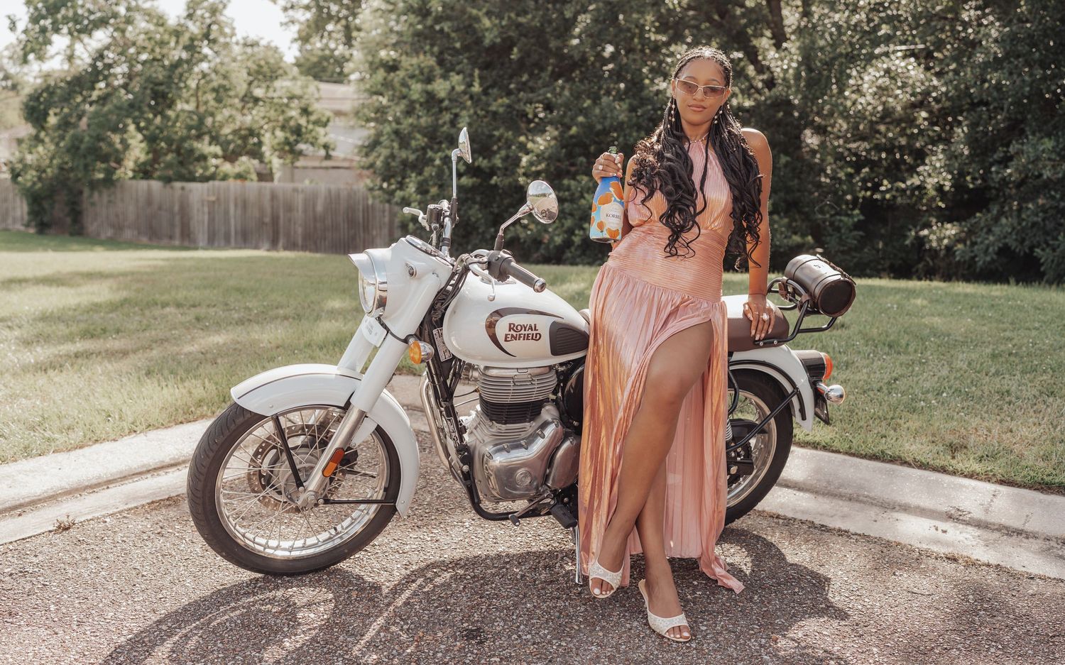 Woman in elegant pink gown posing beside a white Harley-Davidson motorcycle on a sunny day.