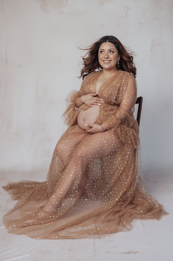 Pregnant woman in a sheer gold glitter maternity gown sitting gracefully in a studio photo session.