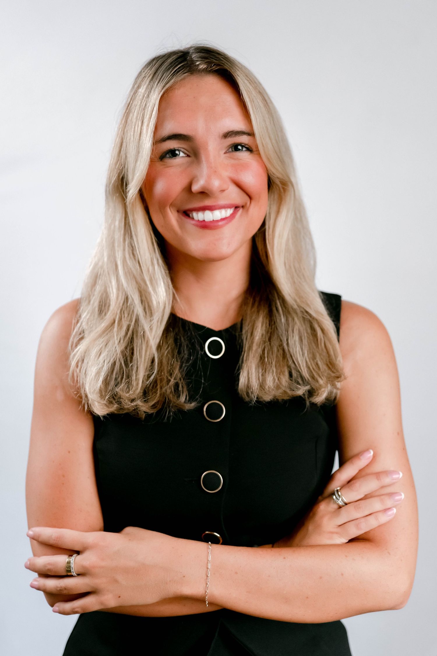 A person with blonde hair wearing a black sleeveless top poses with crossed arms and a bright smile against a white background.
