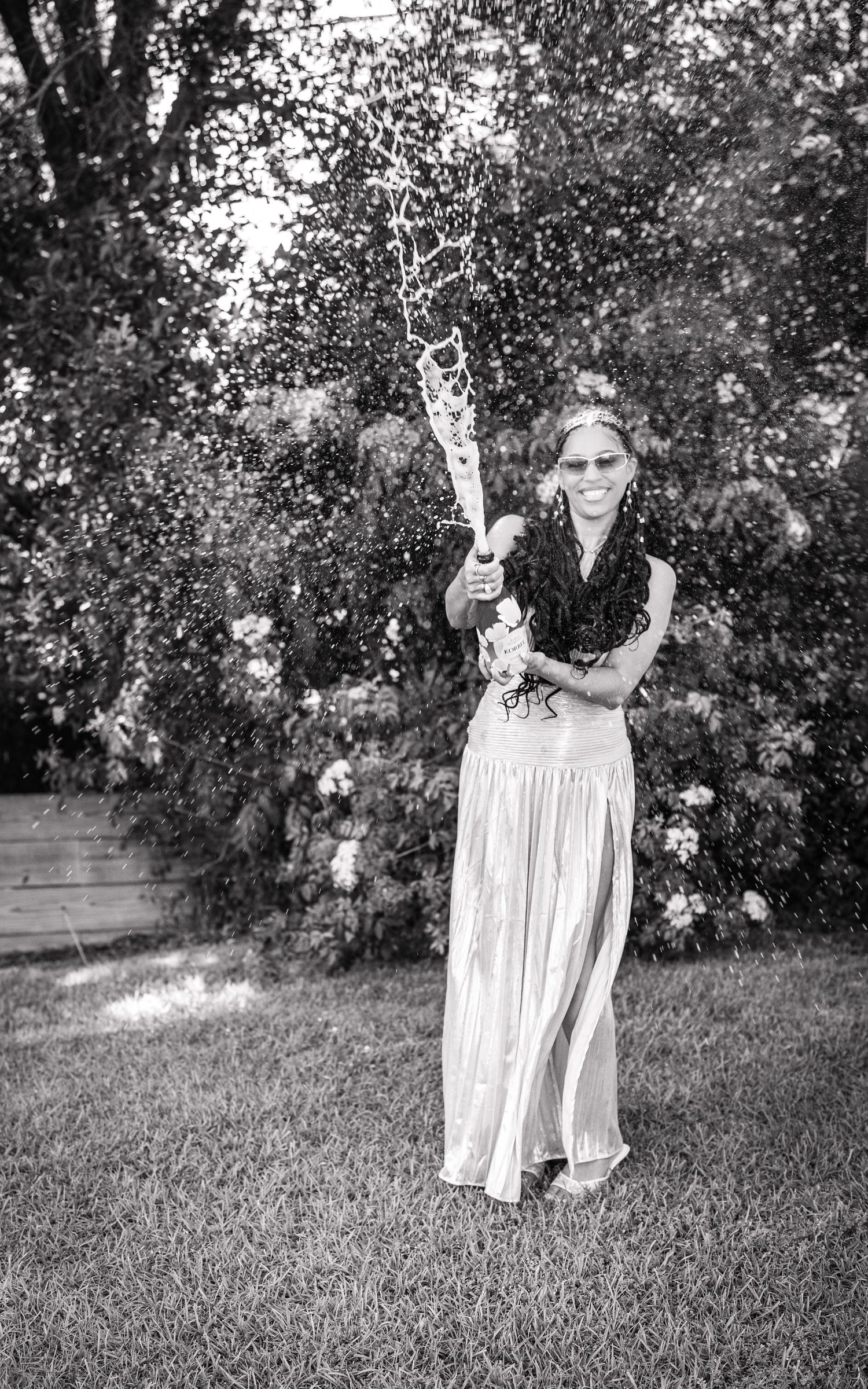 Woman in elegant dress popping champagne bottle outdoors in black and white photo.